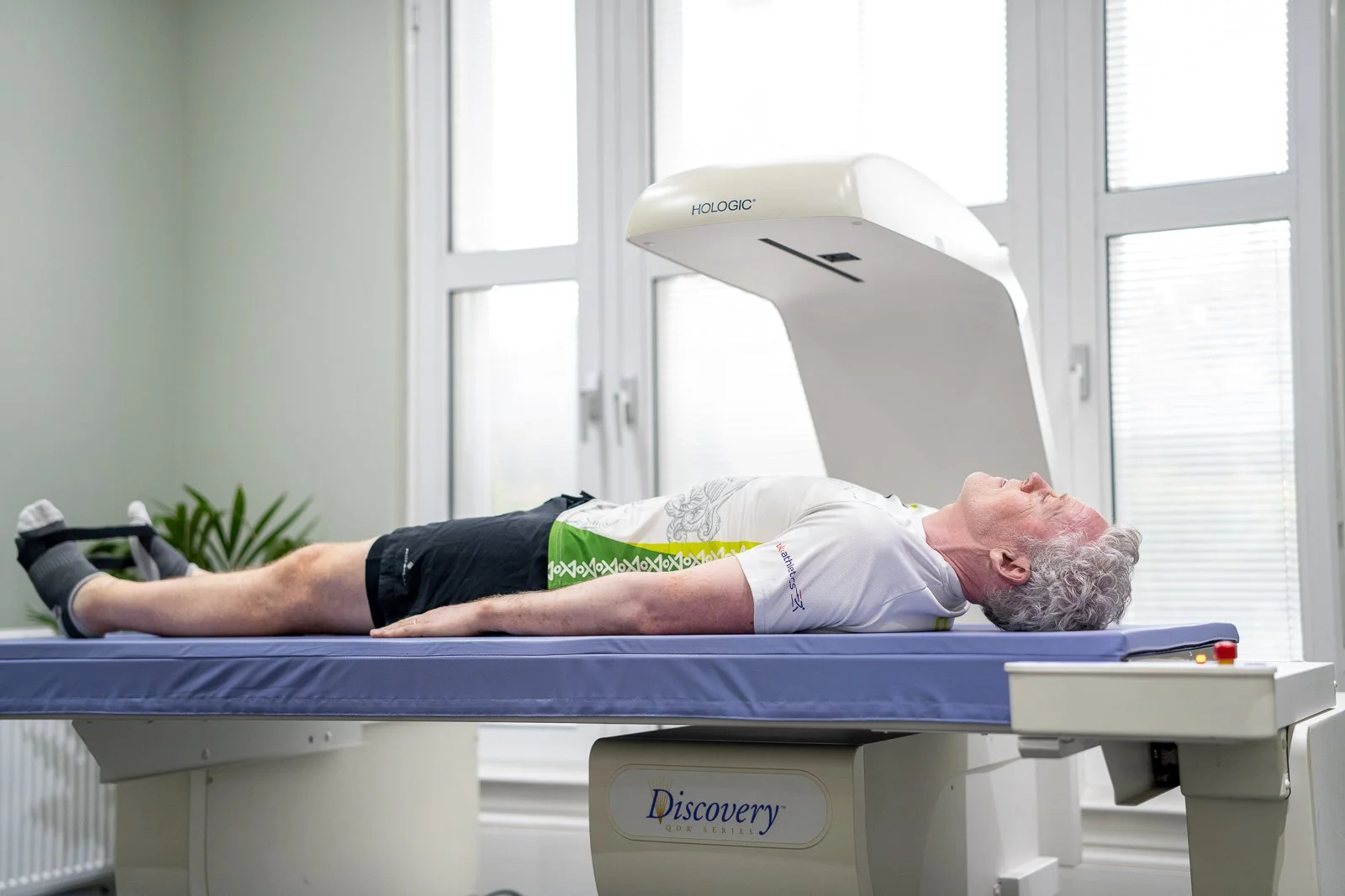 A man lying on his back on a DEXA scanner, smiling and looking up at the ceiling, with a plain wall background.