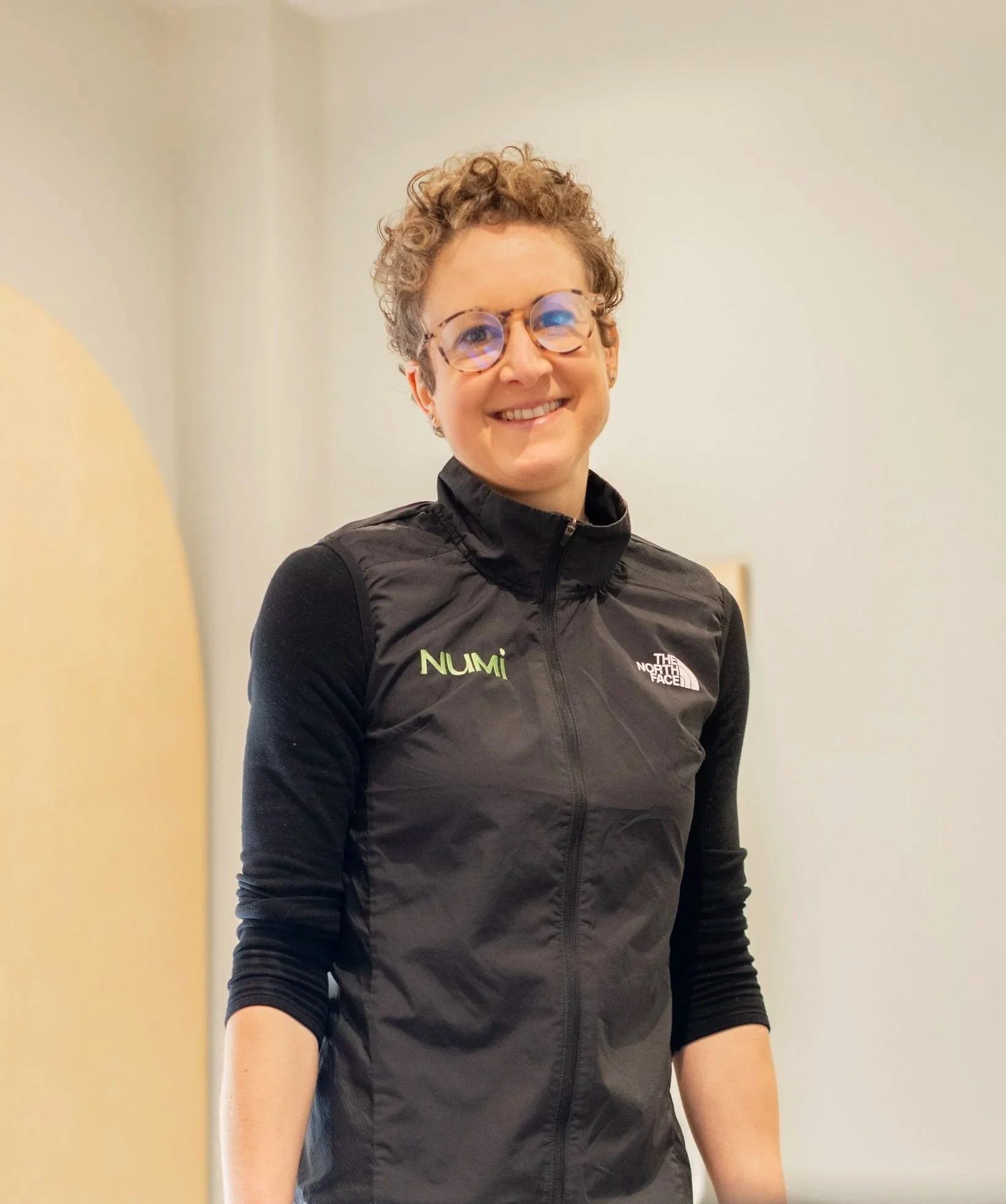 Close-up photo of a smiling woman, Maria the NuMi founder. She has short curly light brown hair and is wearing a black jacket, and standing outside NuMi HQ with wooden shelves in the background.