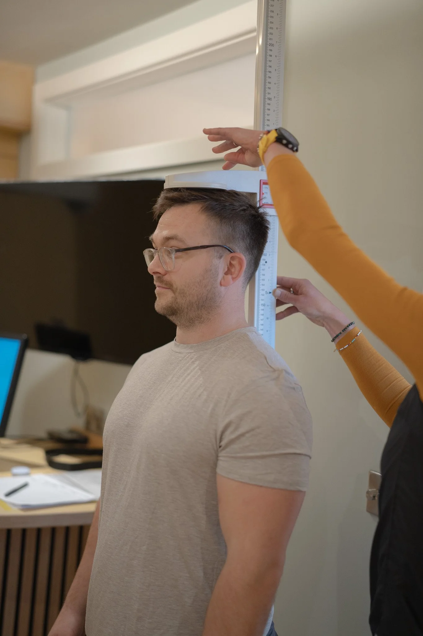 A man wearing glasses stands as a person measures his height using a wall-mounted stadiometer. The person measuring is wearing a yellow long-sleeve shirt and is using a measuring tape attached to the stadiometer.