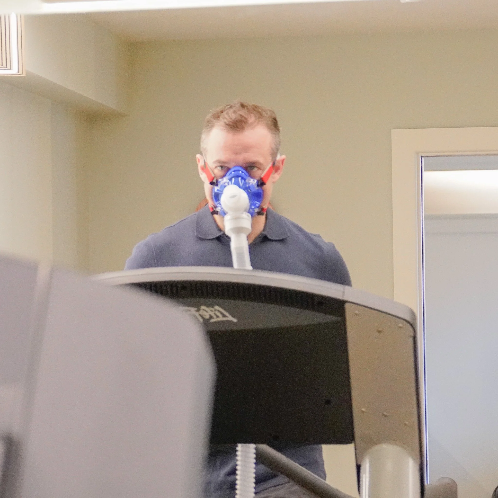 A man wearing a face mask with a breathing tube connected to a machine is having a VO2 max and FatMax assessment, completed by assessing oxygen usage whilst running on a treadmill.