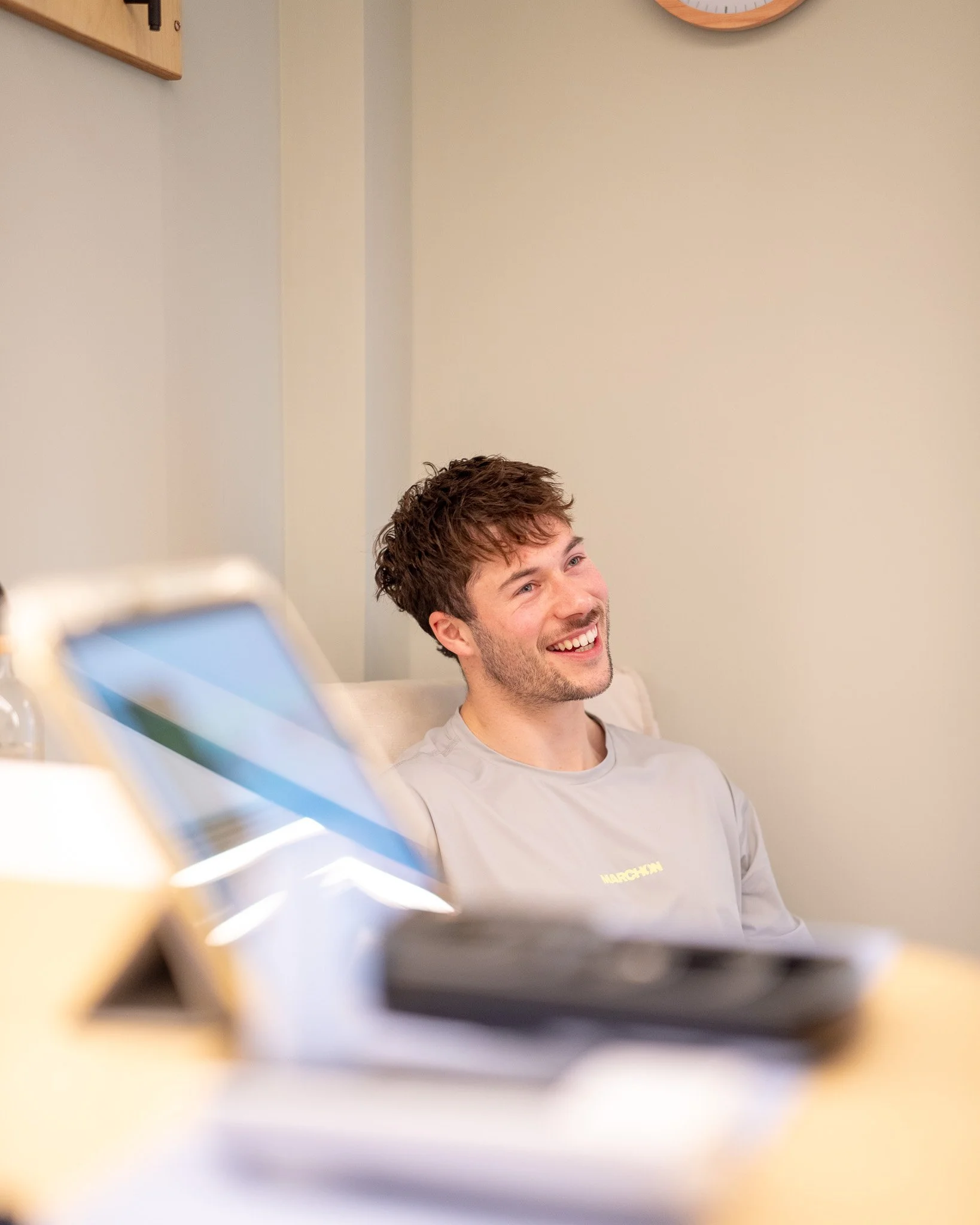 A young man with dark, wavy hair and a beard, wearing a light gray shirt, sitting on a bed or couch, smiling and looking to the side. In the foreground, blurred out, is a tablet or electronic device, with a beige wall and a clock in the background.