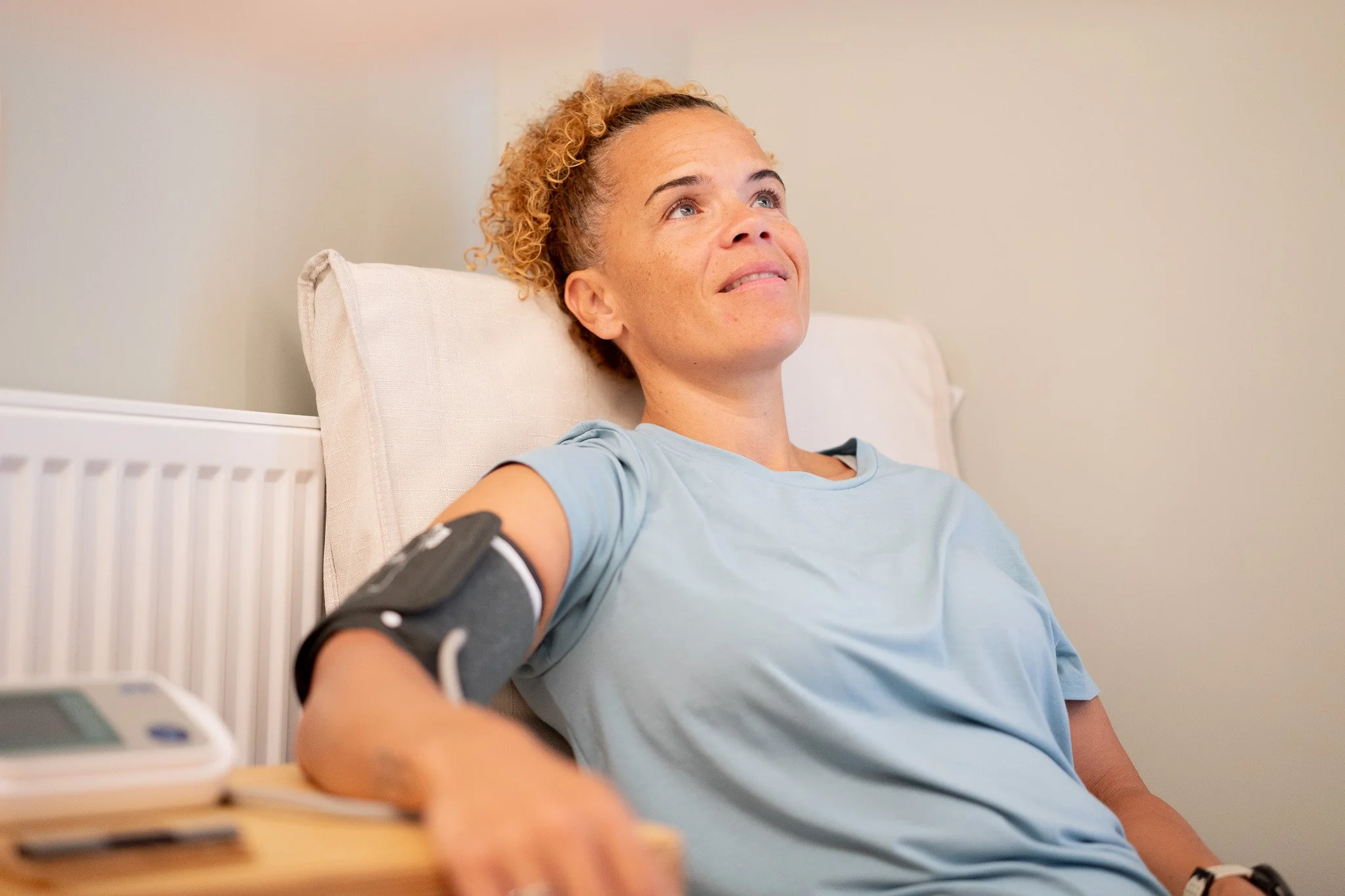 A woman in a light blue shirt is sitting up in a bed, looking slightly upward with a content expression. She has curly blonde hair and a blood pressure cuff on her upper arm, connected to a blood pressure monitor on a bedside table.