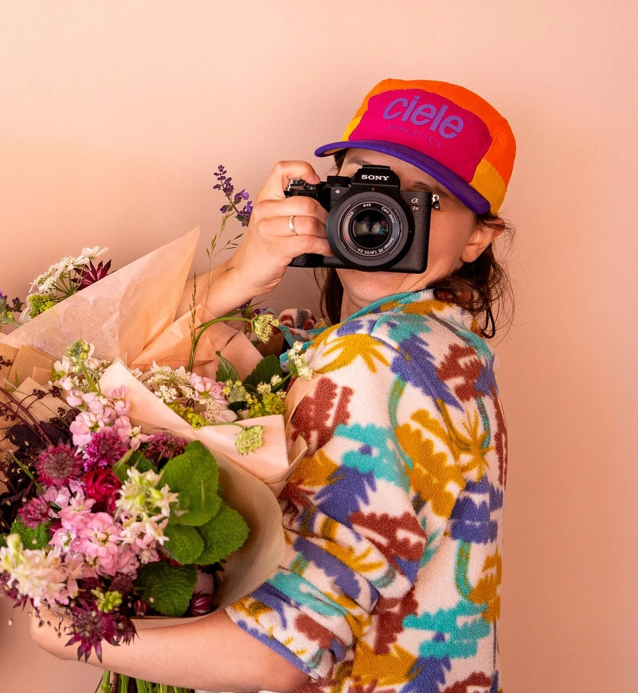 Person holding a camera with a bouquet of flowers, wearing a colorful patterned sweater and a multicolored cap against a pink background.