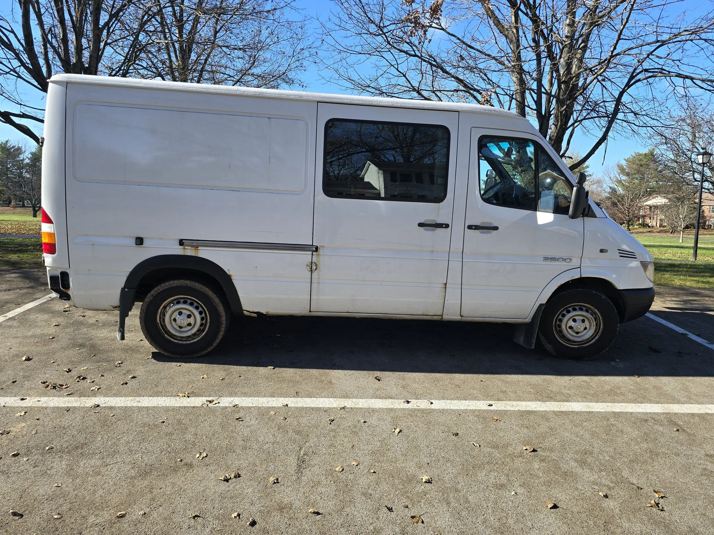 A white cargo van parked in a parking lot during daytime, with trees and a park in the background.