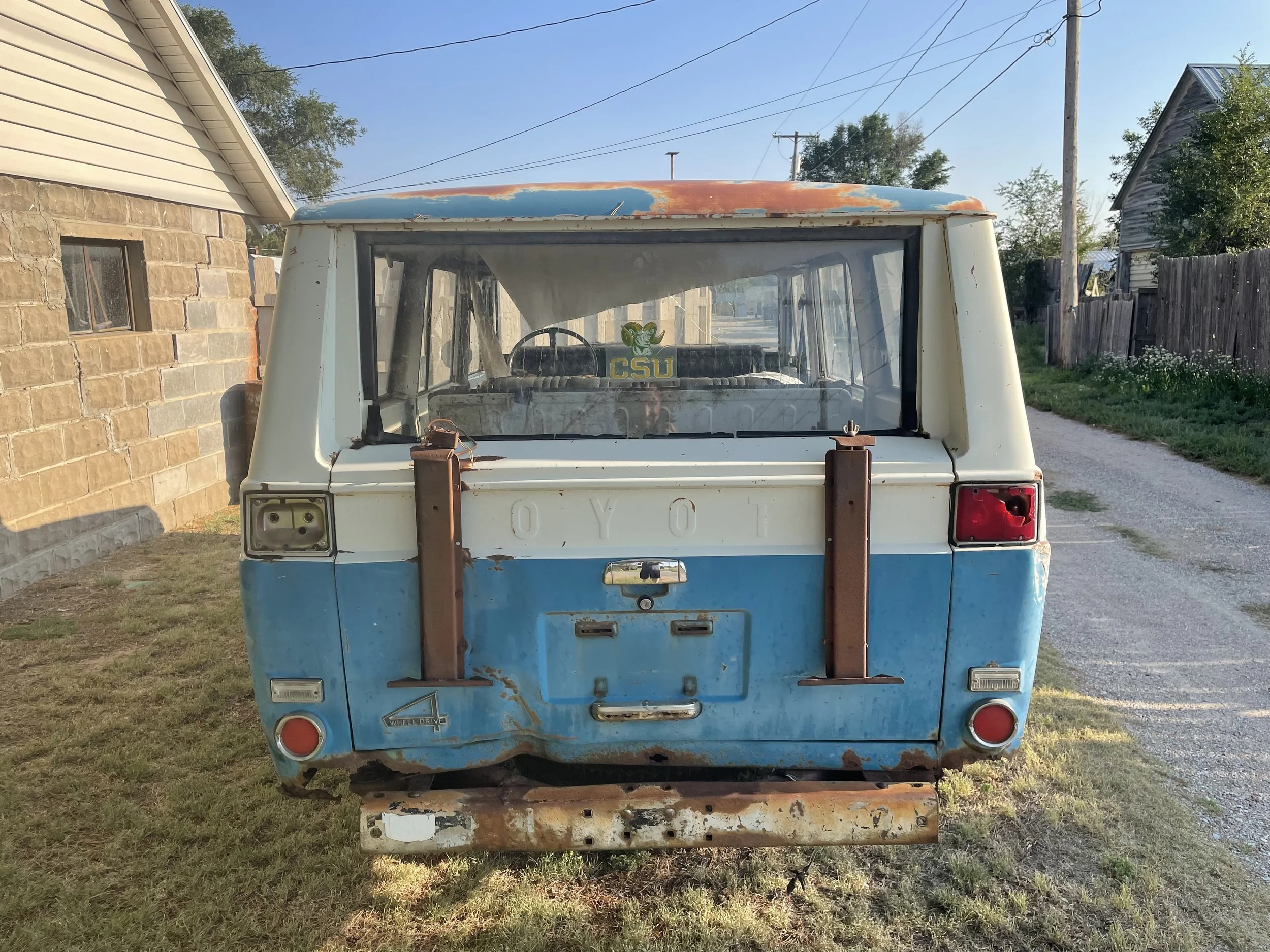 Rear view of an old, rusted Toyota van with a blue and white body, missing rear lights and bumper, parked on grass in front of a brick house and gravel path.