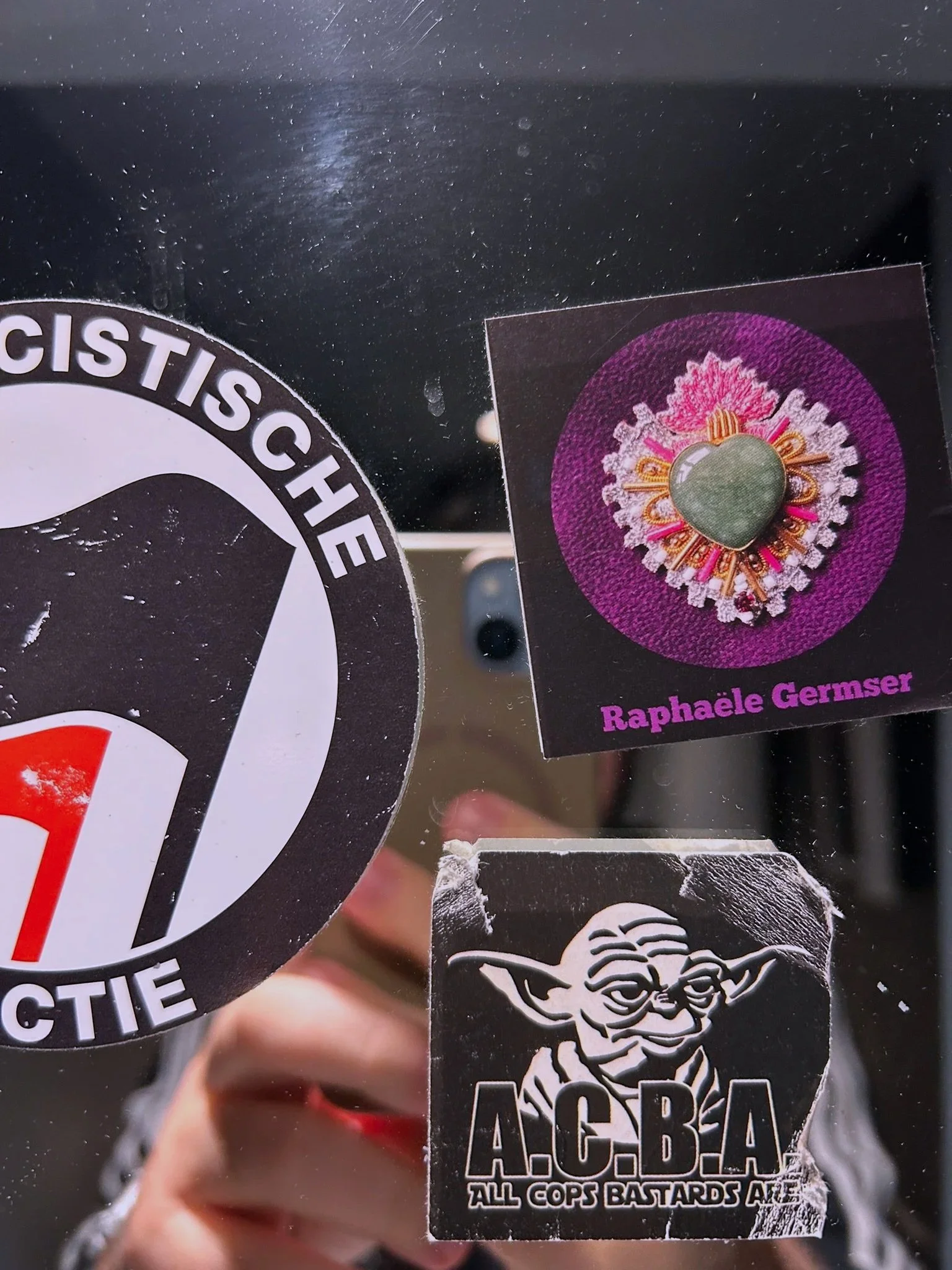 Reflective surface with three stickers: a circular black and white sticker with text, a rectangular sticker with a heart-shaped green gemstone brooch against a purple background and the name Raphaèle Germser, and a square sticker with an illustration of Yoda and the text 'A.C.B.A. All Cops Bastards Are'.