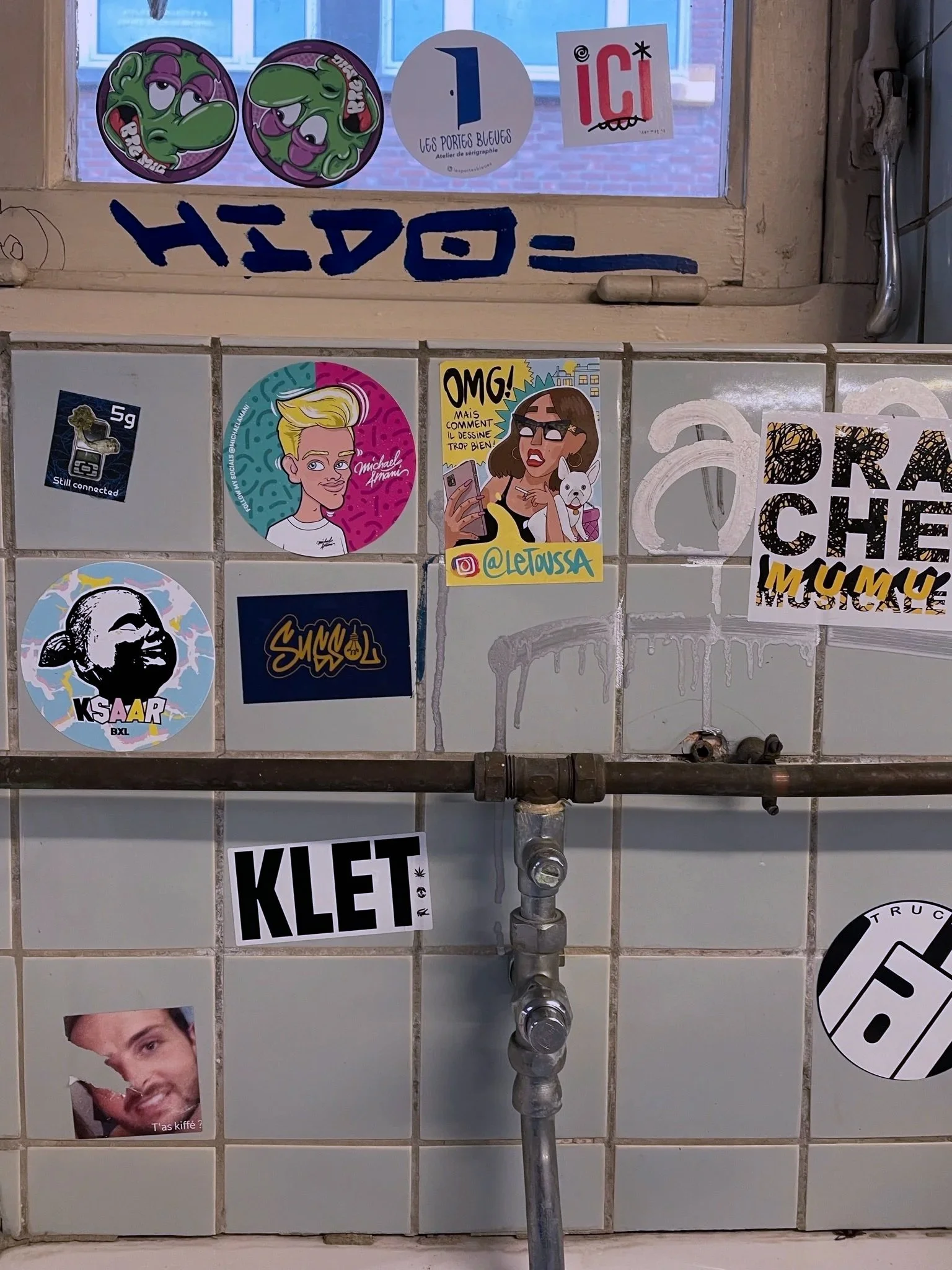 A tiled wall with various stickers and graffiti, including cartoon characters, text, and graphic designs.