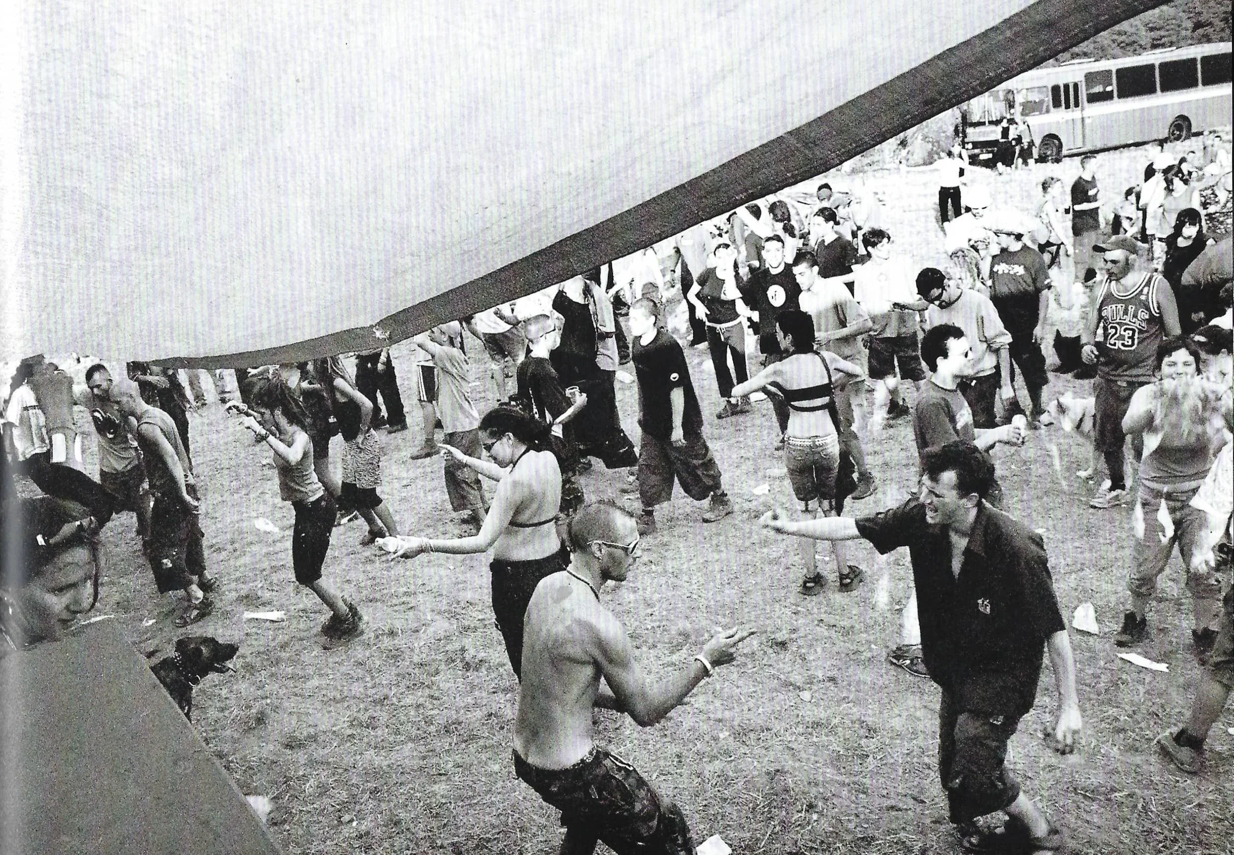 Black and white photo of a large group of people dancing together outdoors, with some wearing masks and casual clothing, in front of a bus.