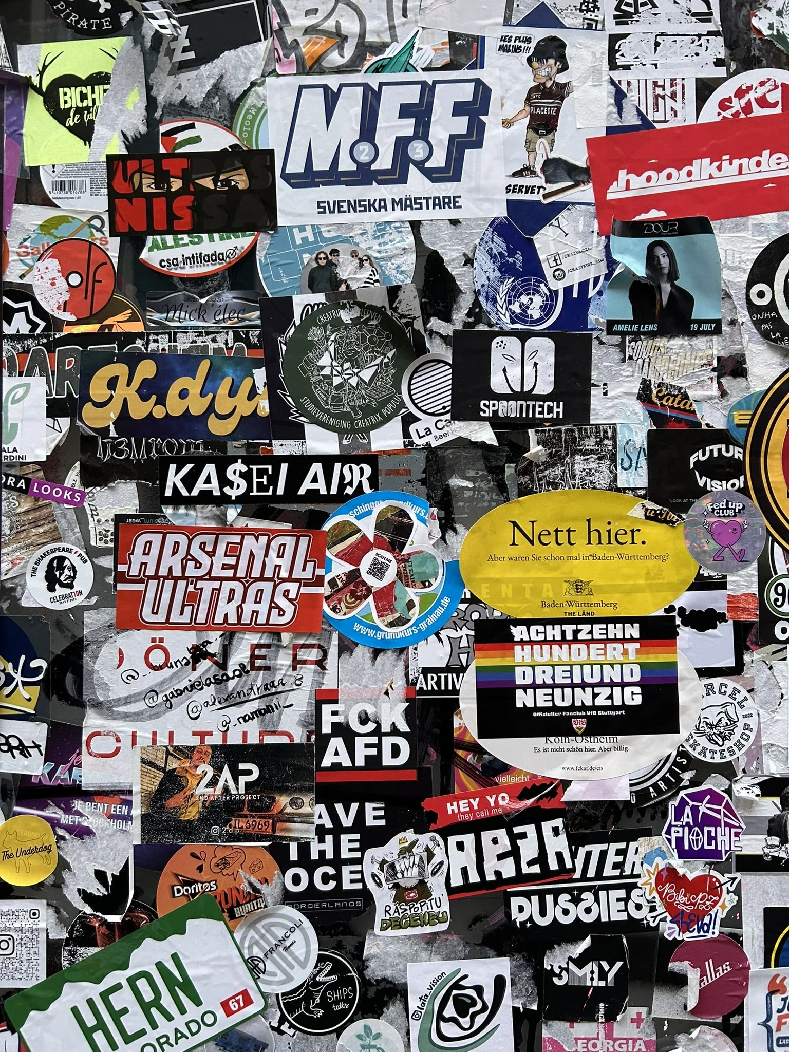 A collection of colorful stickers pasted on a surface, featuring various texts, logos, and graphics, including stickers with political slogans, brand logos, and artistic designs.