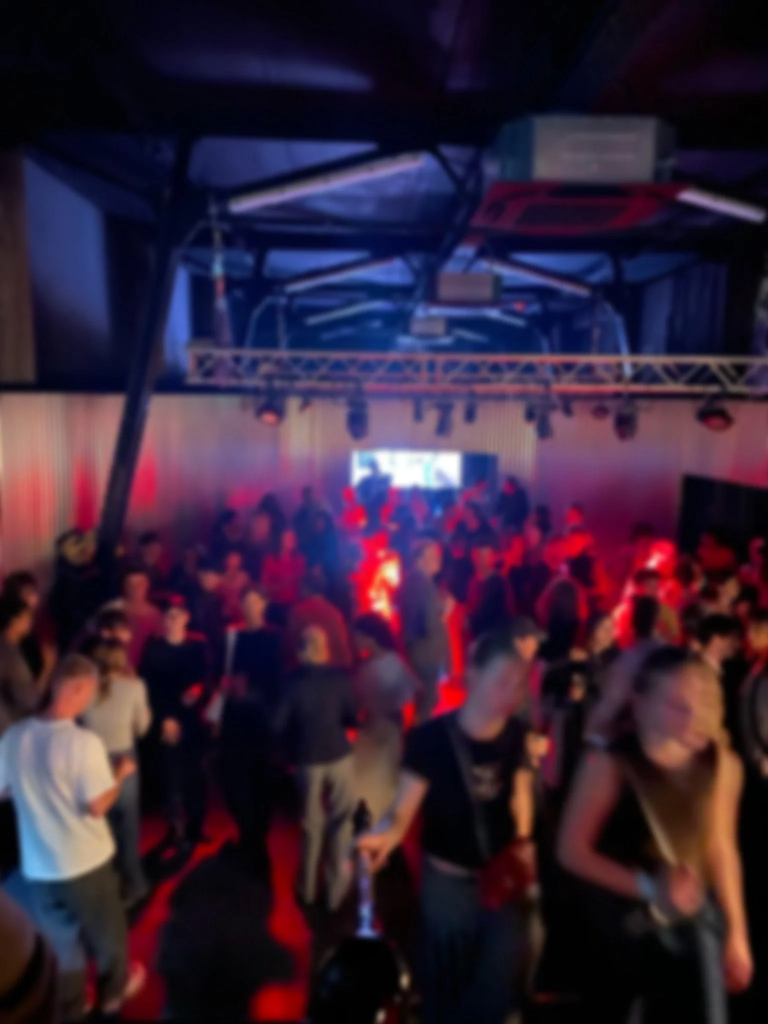 A crowded nightclub with people dancing and socializing under colorful red and purple lighting, with a DJ booth and screen at the front.