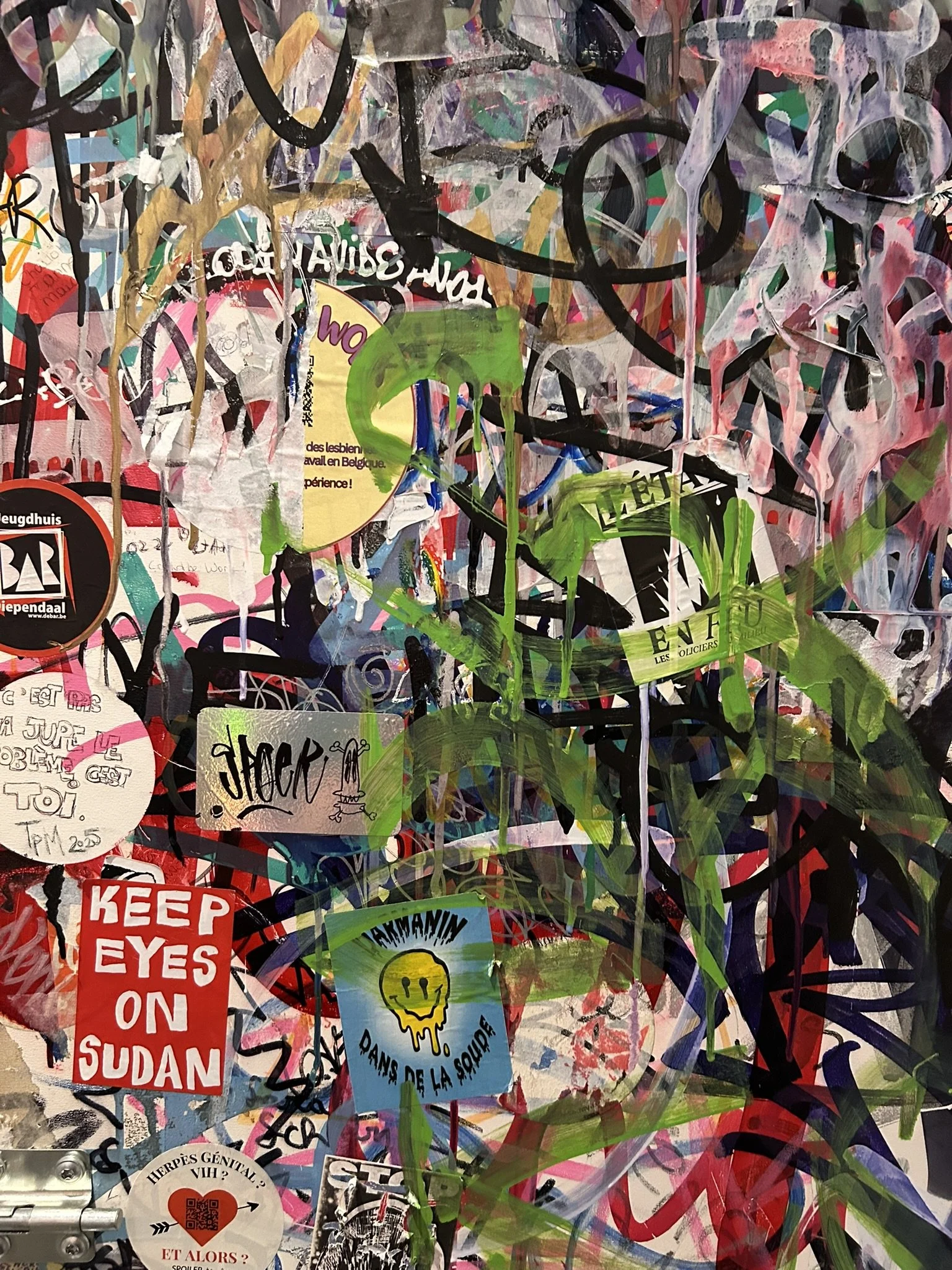 A densely covered wall with various overlapping graffiti, stickers, and paint splashes in multiple colors, including black, pink, green, white, yellow, and red.
