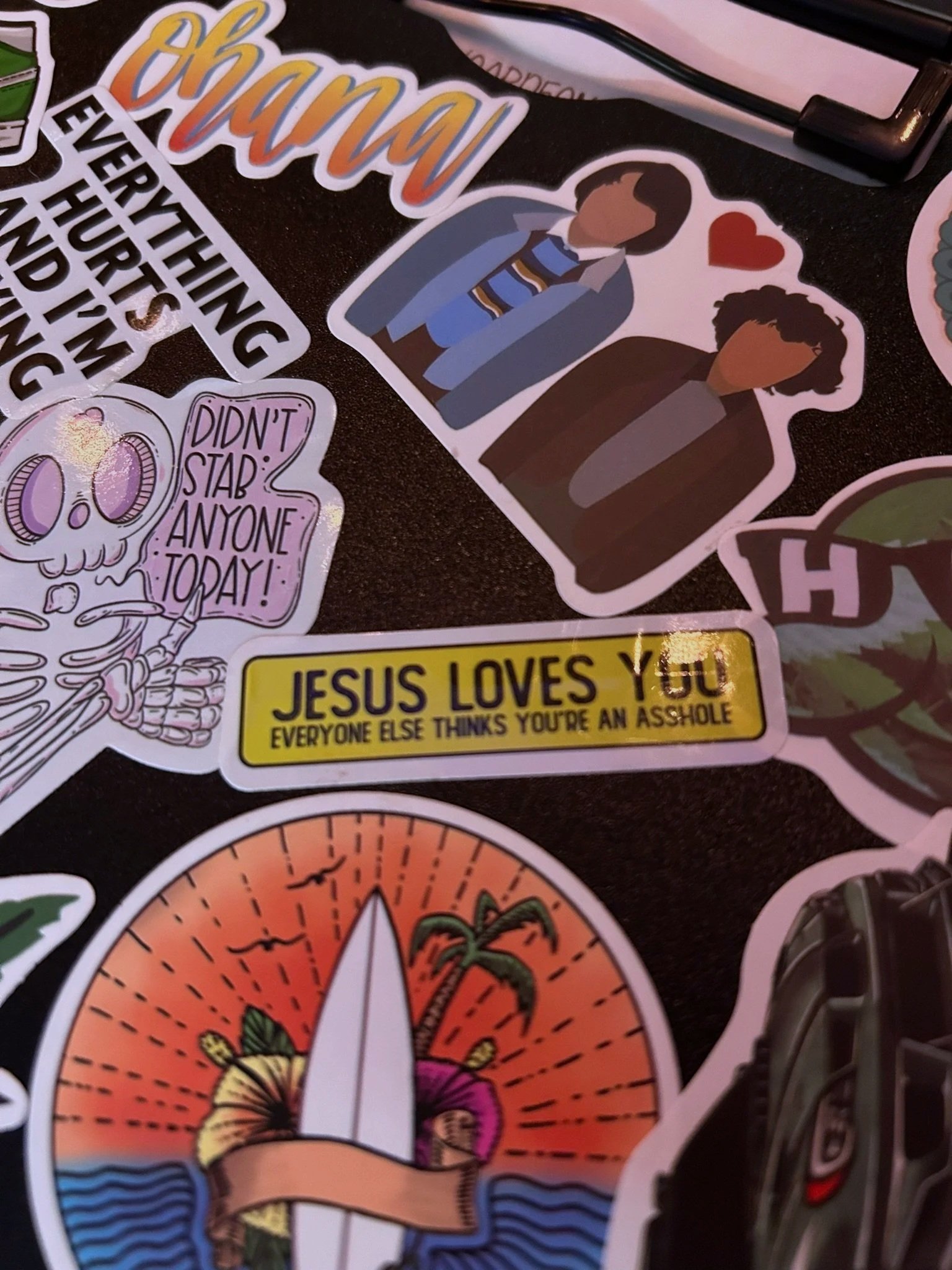 Collection of various stickers including a stylized cartoon of two people with a red heart above, a rainbow-colored sunrise with a surfboard and a banner, a skeleton holding a sign that says 'Didn’t stab anyone today!', a text sticker that says 'Jesus loves you everyone else thinks you’re an asshole', and a partial view of a camera.