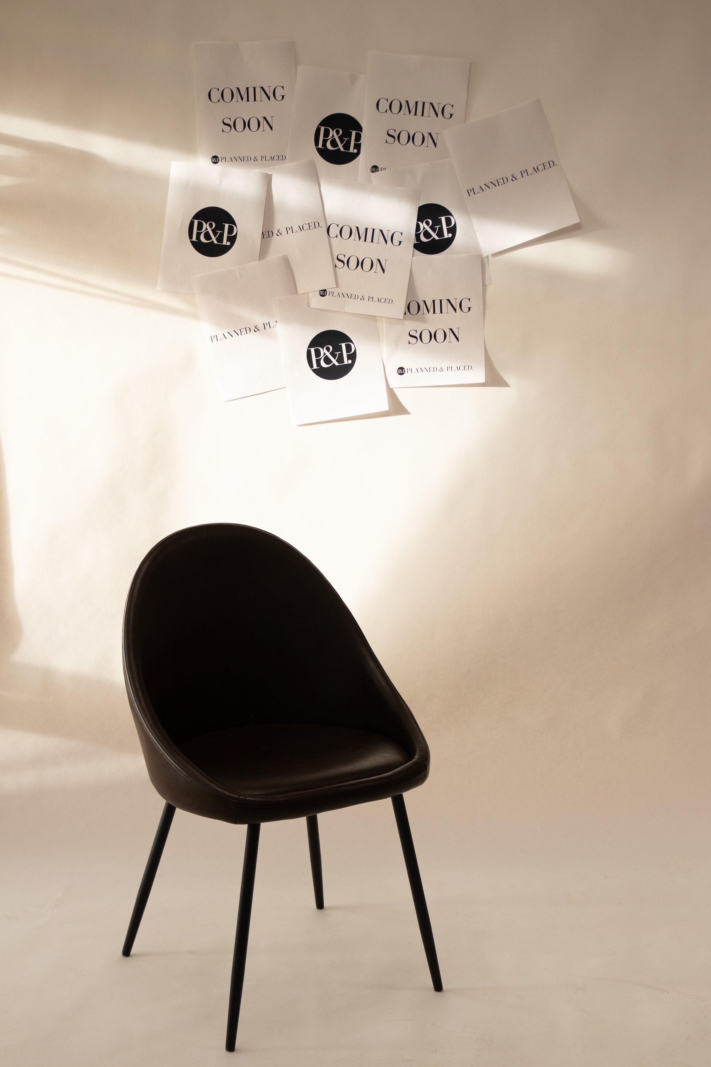 A black modern chair with four thin legs and a rounded seat against a plain wall with multiple signs that read "Coming Soon" and "Planned & Placed," along with a circular logo with "P&P" in the center.