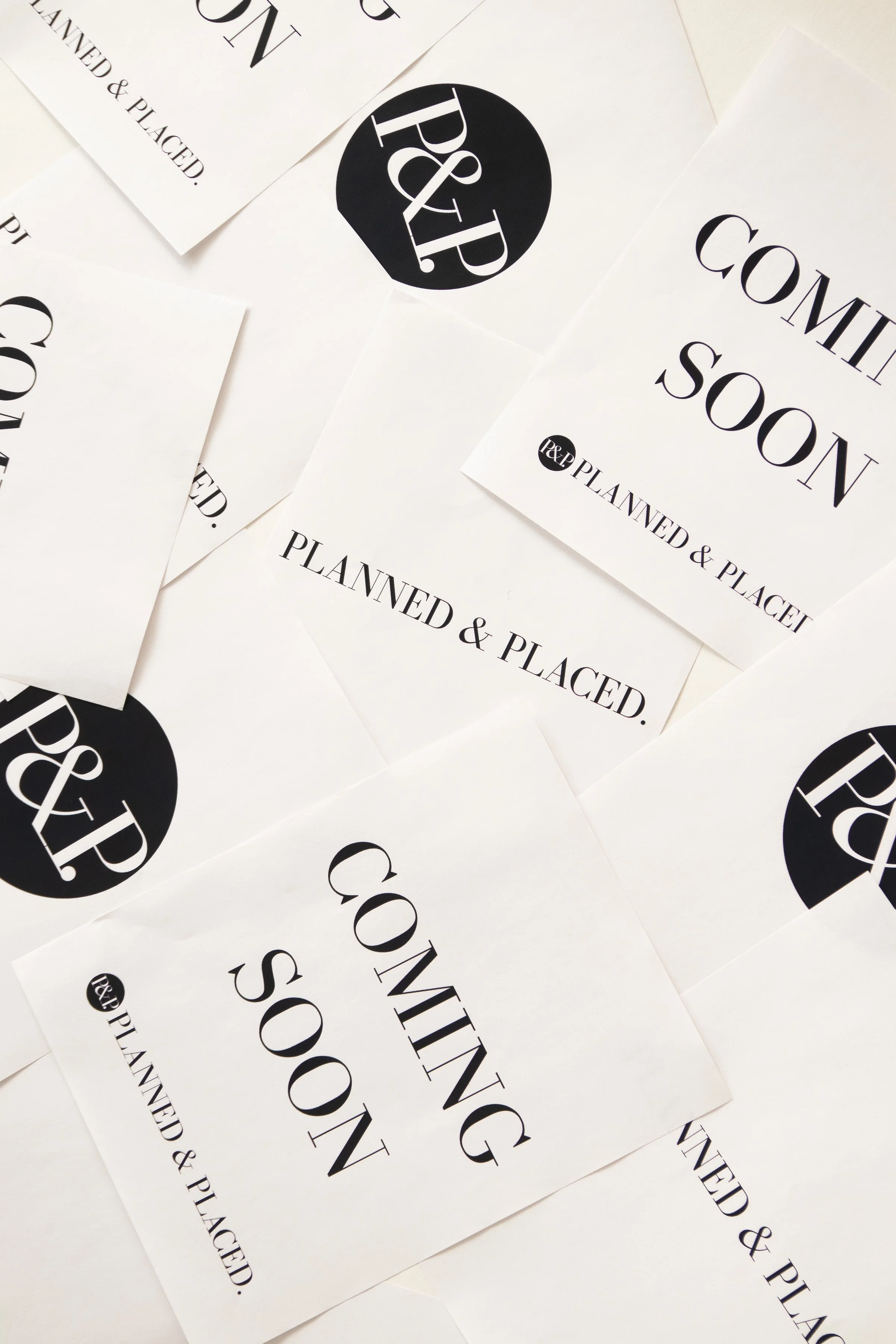 Multiple white sheets of paper with black text and logos, including phrases like 'COMING SOON' and 'PLANNED & PLACED', and a black circle with white initials '& P' over a black circle background.