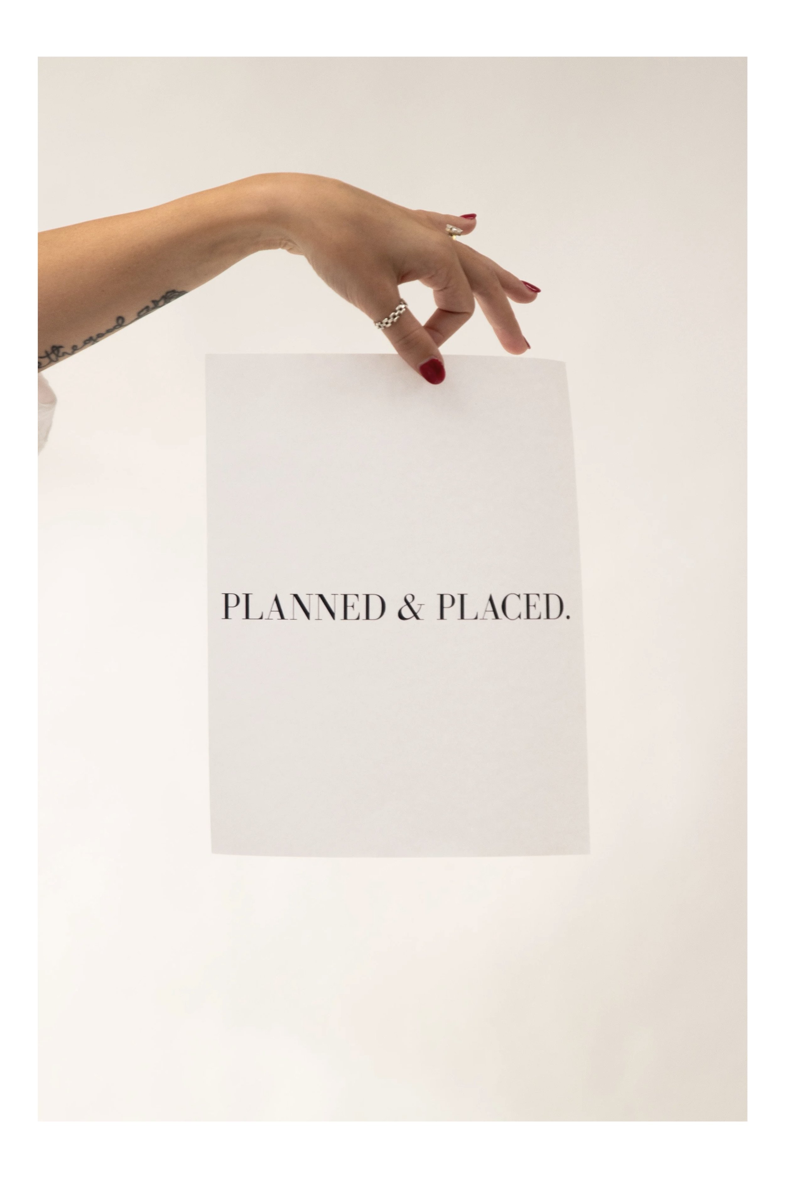A hand with red nails and rings holding a white sheet of paper that reads "PLANNED & PLACED." against a plain white background.