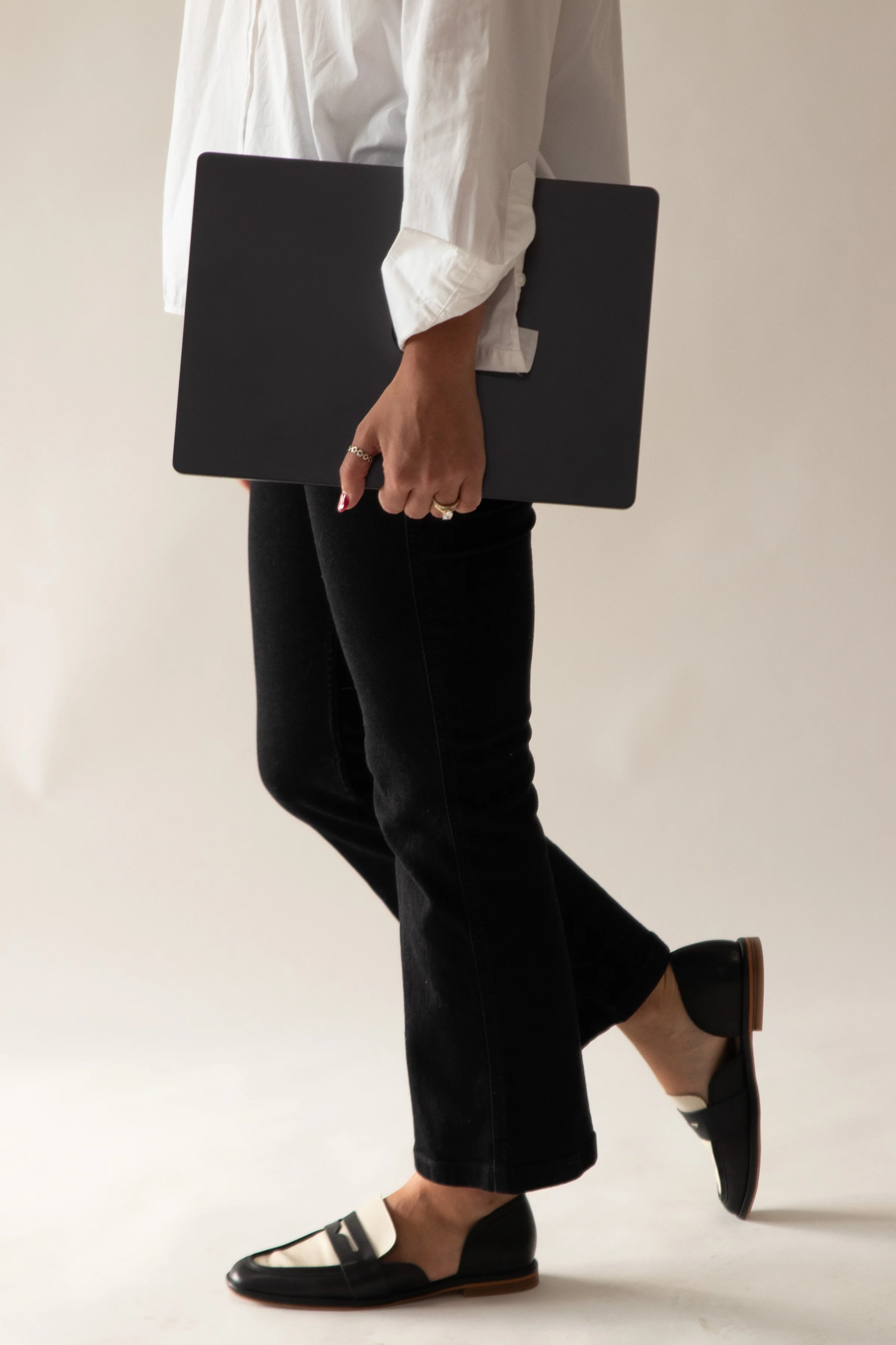 Person holding a closed laptop while walking, wearing a white shirt, black pants, and black and white flats.