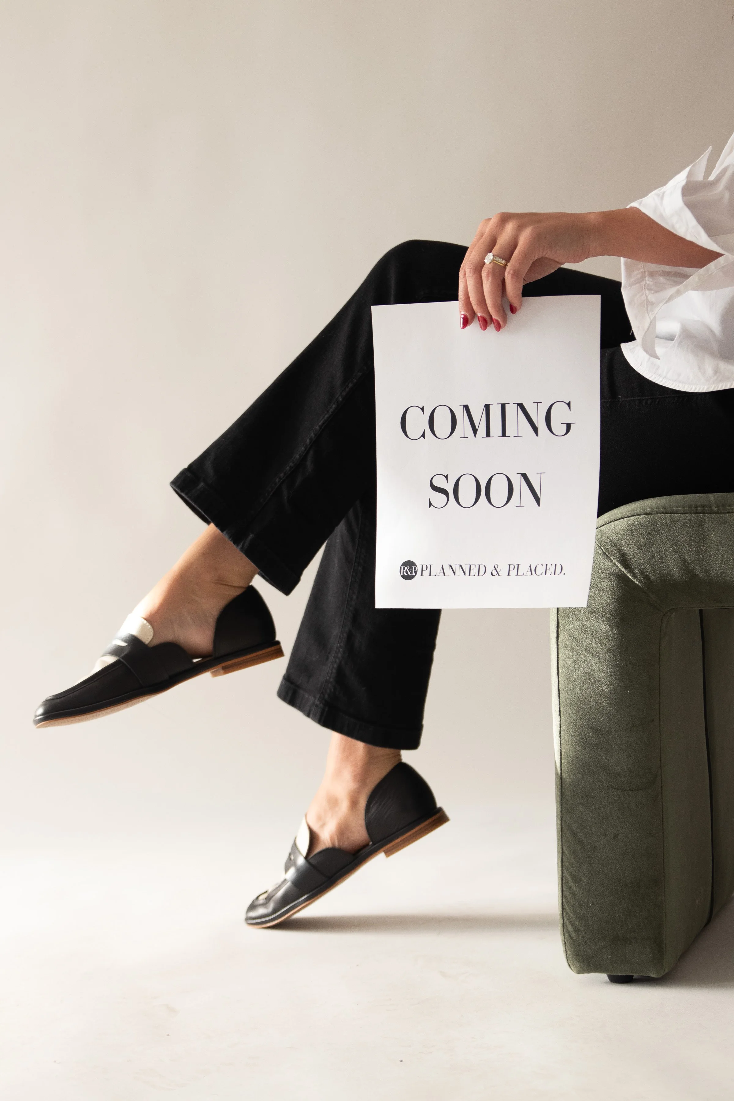 Person sitting on a green couch holding a 'Coming Soon' sign with a white shirt, black pants, and black slip-on shoes, against a plain background.