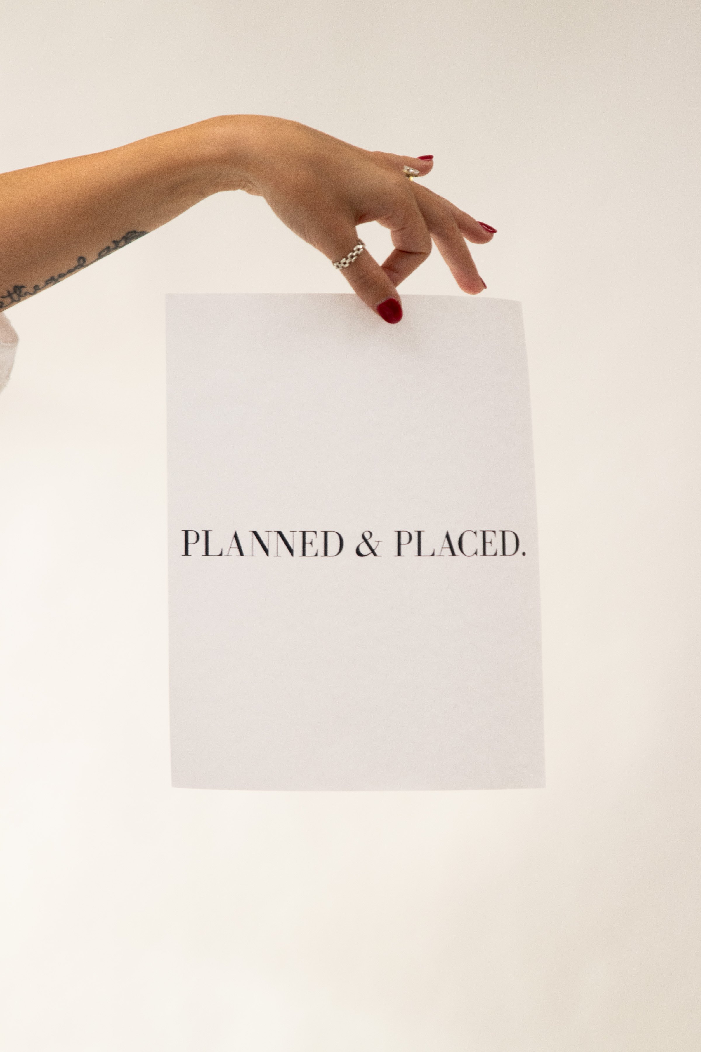 A person's hand holding a piece of paper with the words 'PLANNED & PLACED.' printed on it. The person has red nail polish, rings on fingers, and a tattoo on the wrist.
