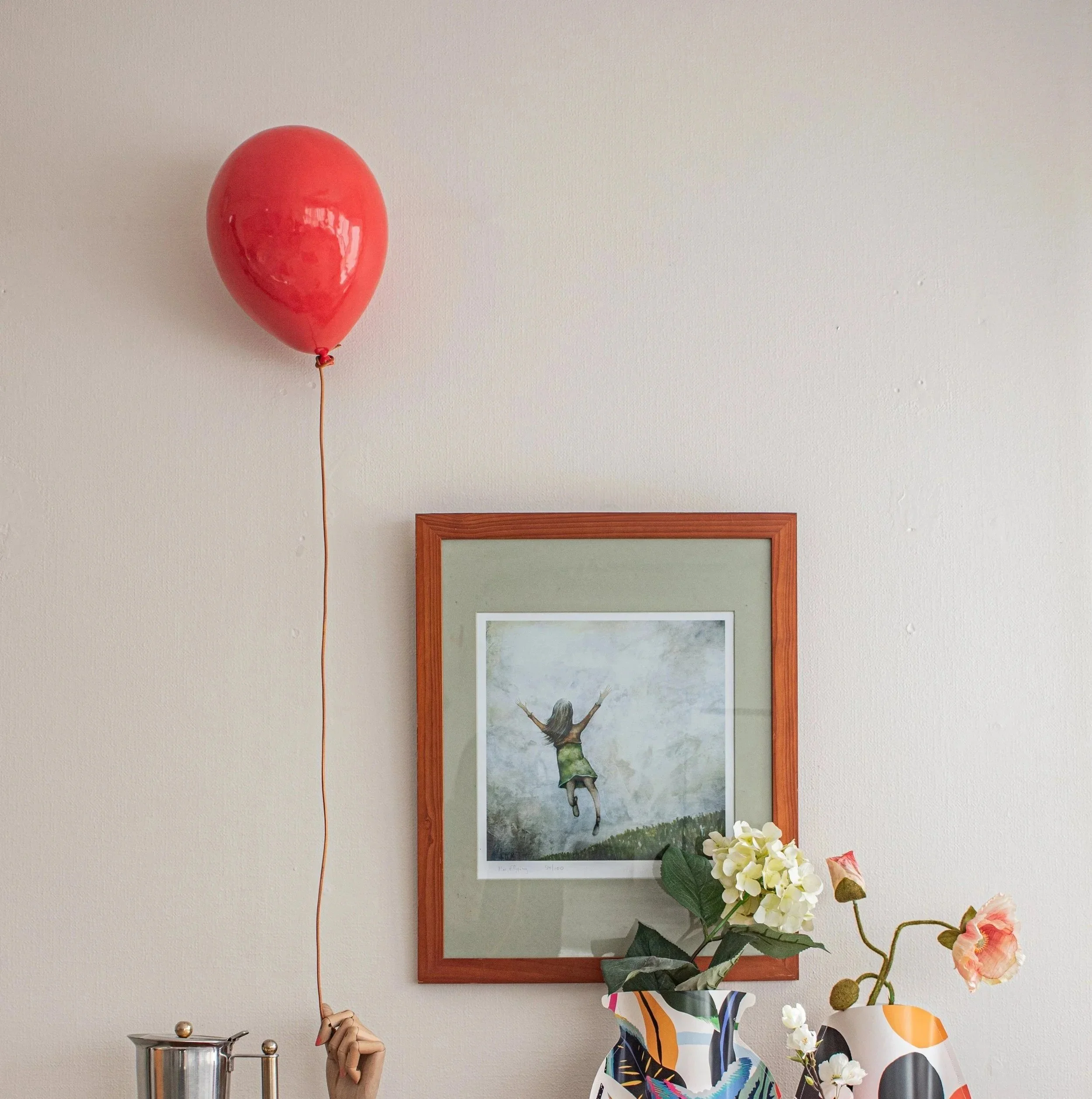 A red balloon attached to a string being held by a hand, with a framed painting of a girl jumping in the air on a wall, and a vase with white and pink flowers on a table.