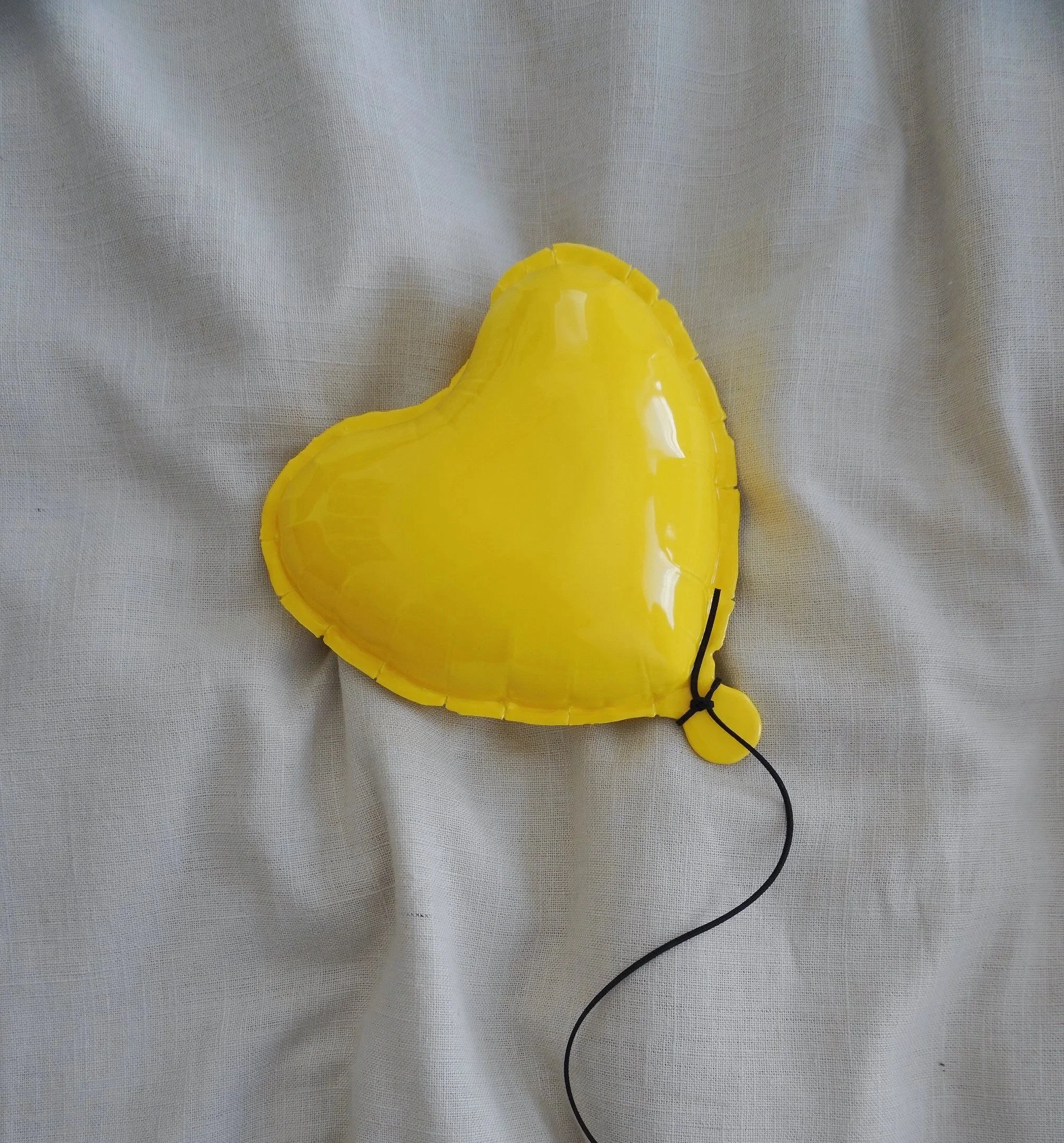 Yellow heart-shaped balloon with a black string, lying on a light-colored fabric surface.