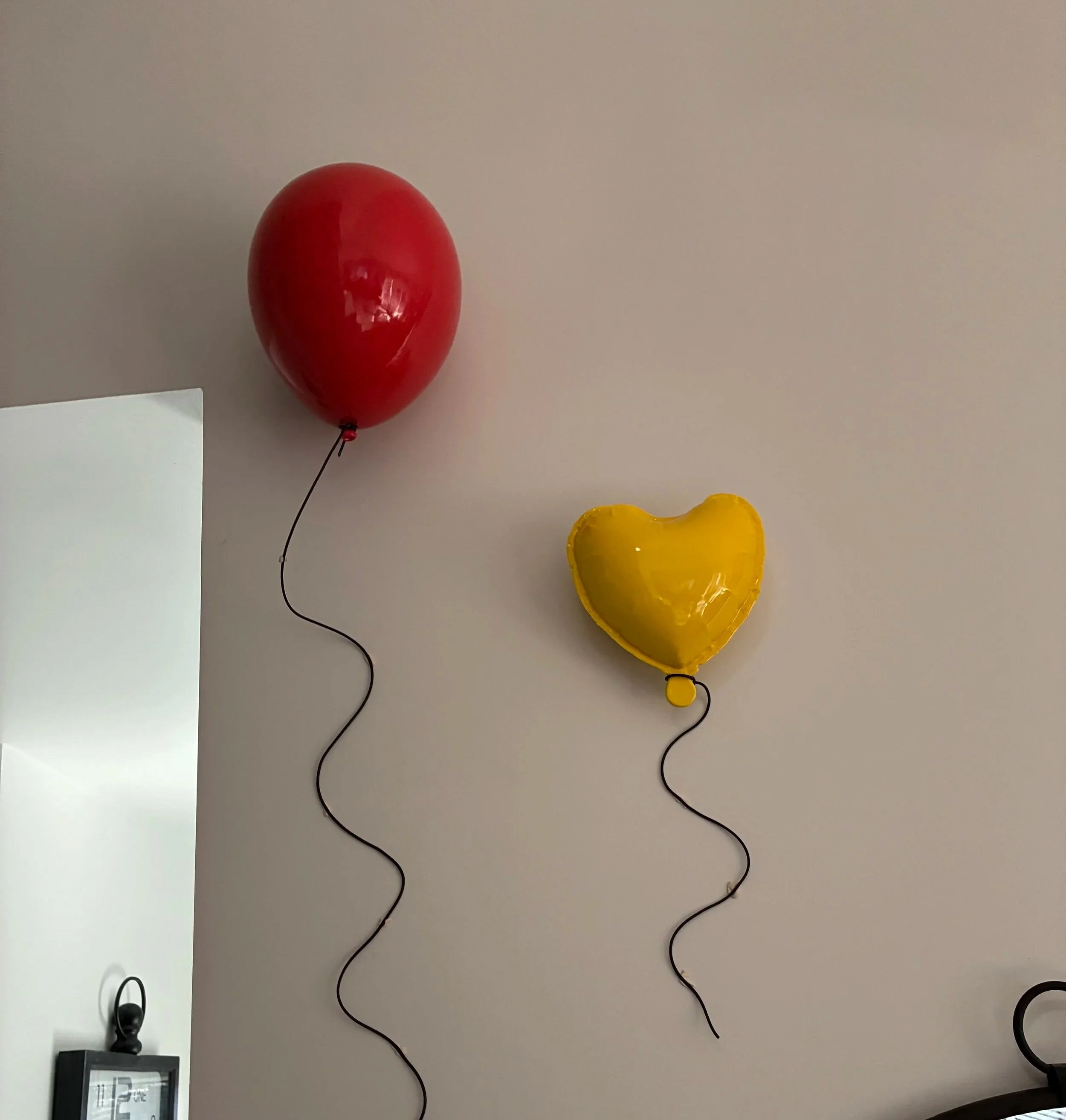 A red balloon and a yellow heart-shaped balloon hanging on a plain wall, with the balloons attached to black strings.