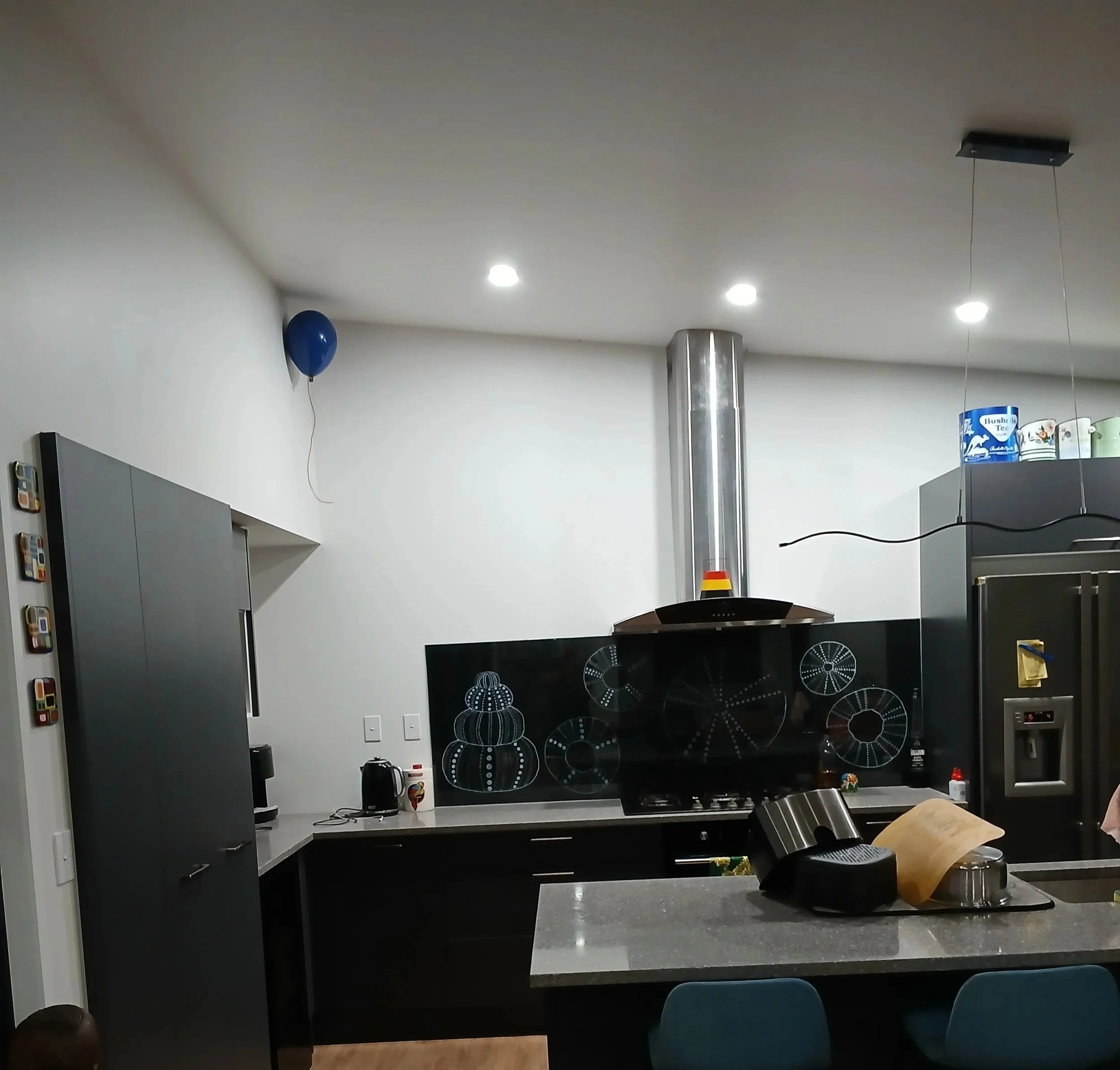 Modern kitchen with black cabinetry, a gray countertop, a stainless steel range hood, and decorative black backsplash with white drawings, a refrigerator, and a balloon on the ceiling.