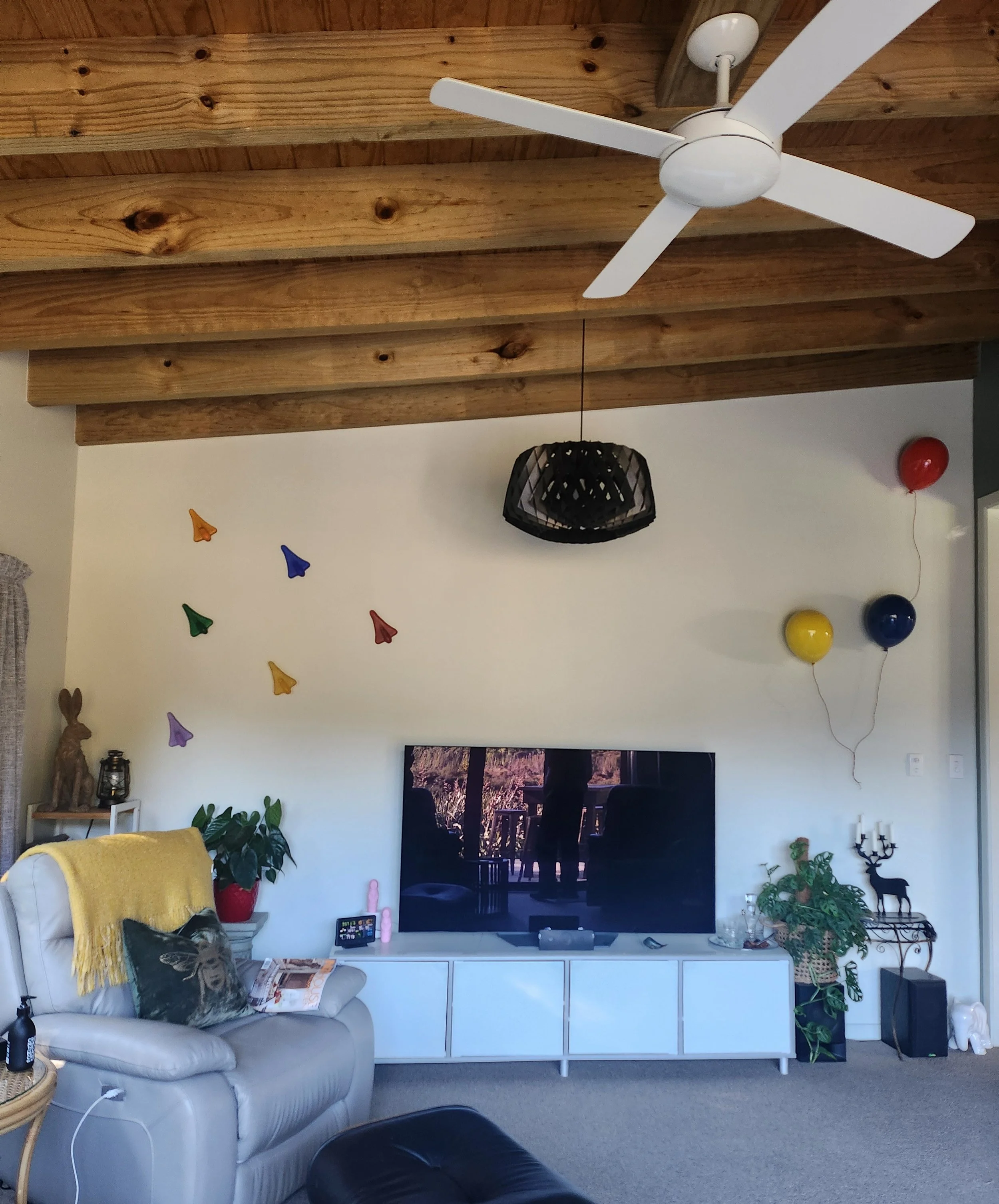 Living room with a white ceiling fan, black pendant light, machine-pressed colorful paper cone decorations on the wall, a entertainment center with a television, a beige armchair with a yellow blanket and a cat pillow, houseplants, balloons, and vari