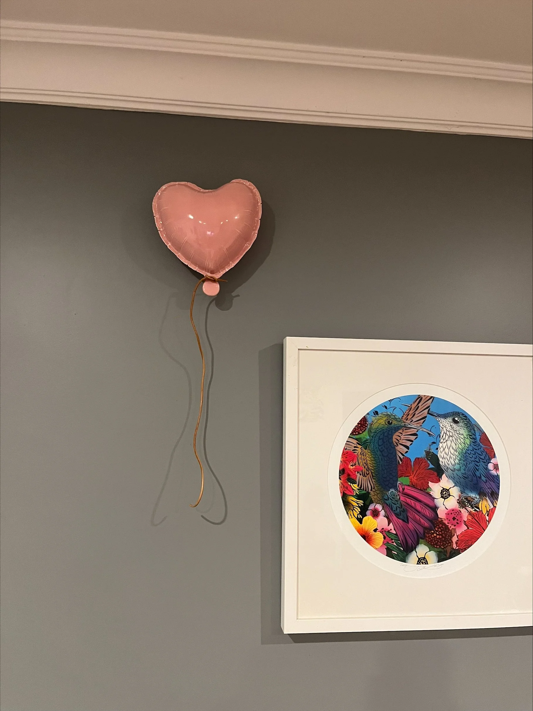 Pink heart-shaped balloon hanging on a gray wall next to framed artwork of two hummingbirds surrounded by colorful flowers.