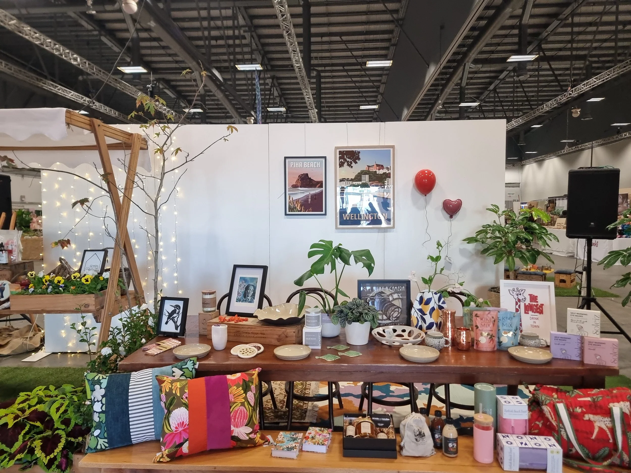 An indoor display booth decorated with plants, picture frames, and various items including ceramics, books, and decorative objects. There are balloons, framed photos of beach scenes, and small light strings on a white wall in the background.