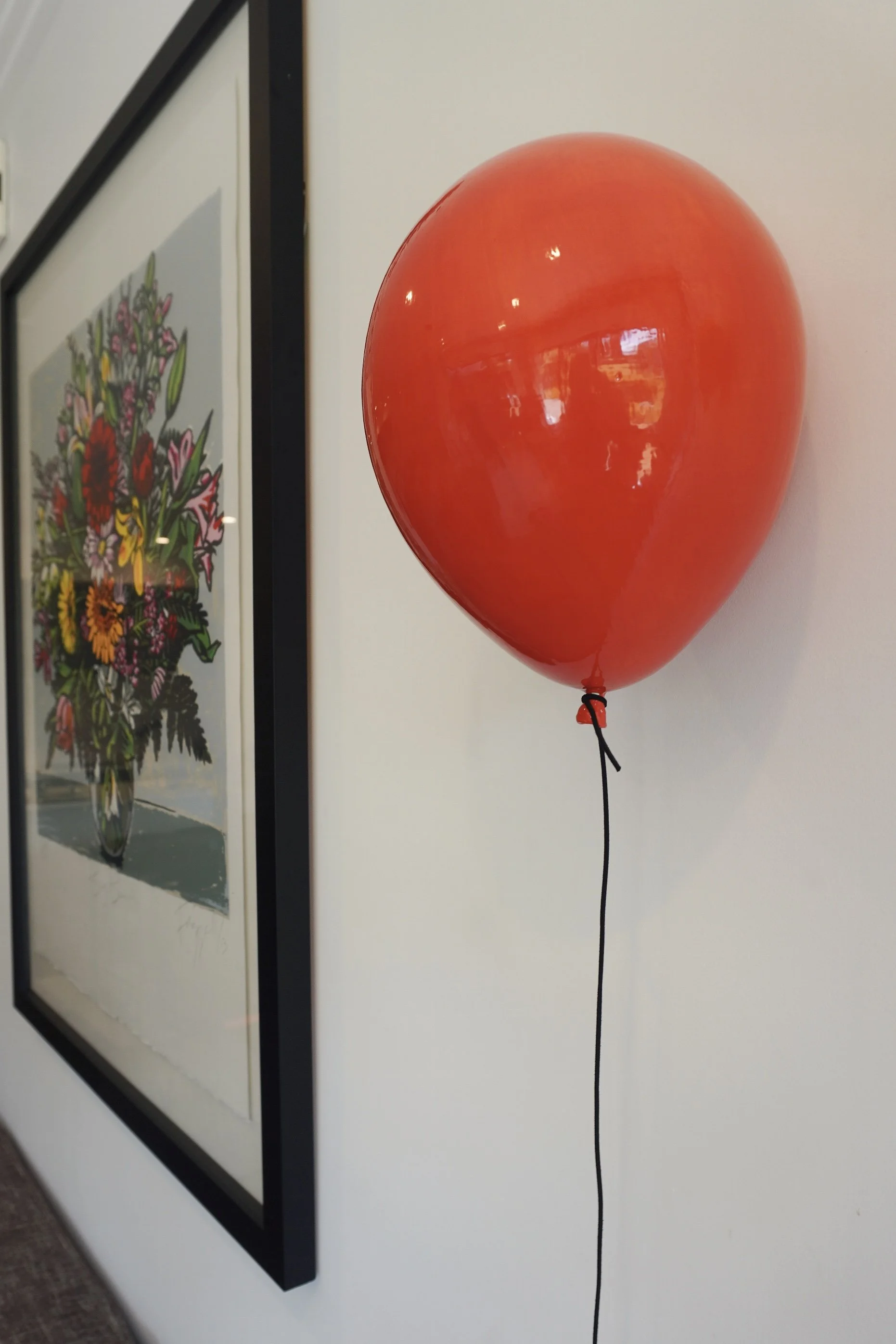 Red helium balloon tied to a black string hanging on a wall, with framed floral artwork in the background.
