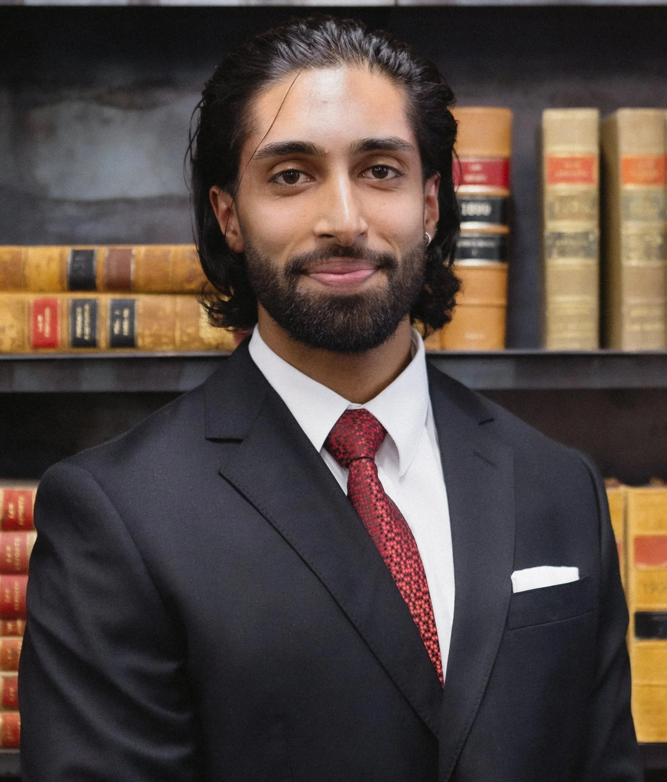 A man in a black suit, white shirt, and red tie standing in front of a bookshelf filled with old books.