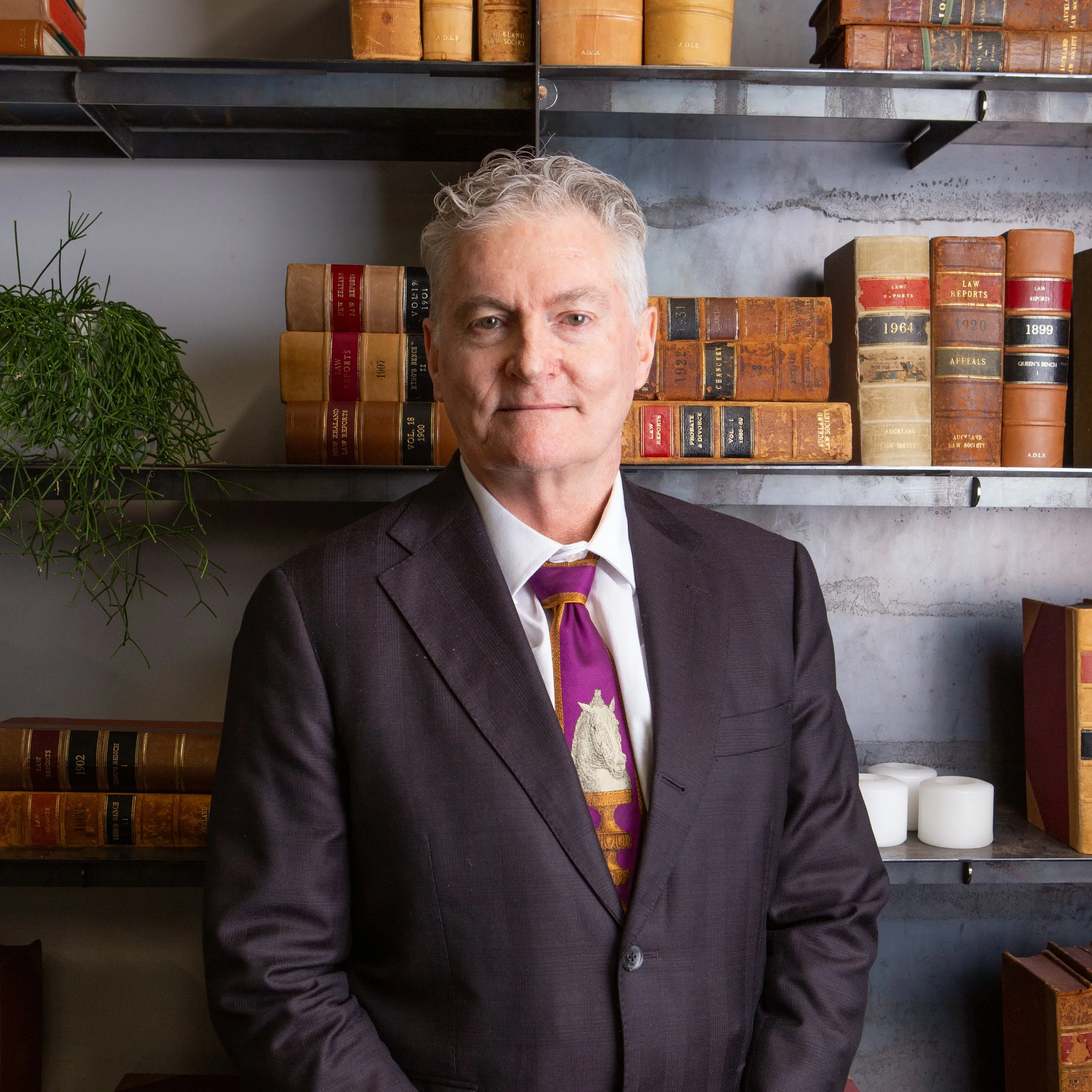 A man with gray hair, wearing a dark suit and a white shirt, stands in front of a metal shelf filled with old law books and a small green plant on the left.
