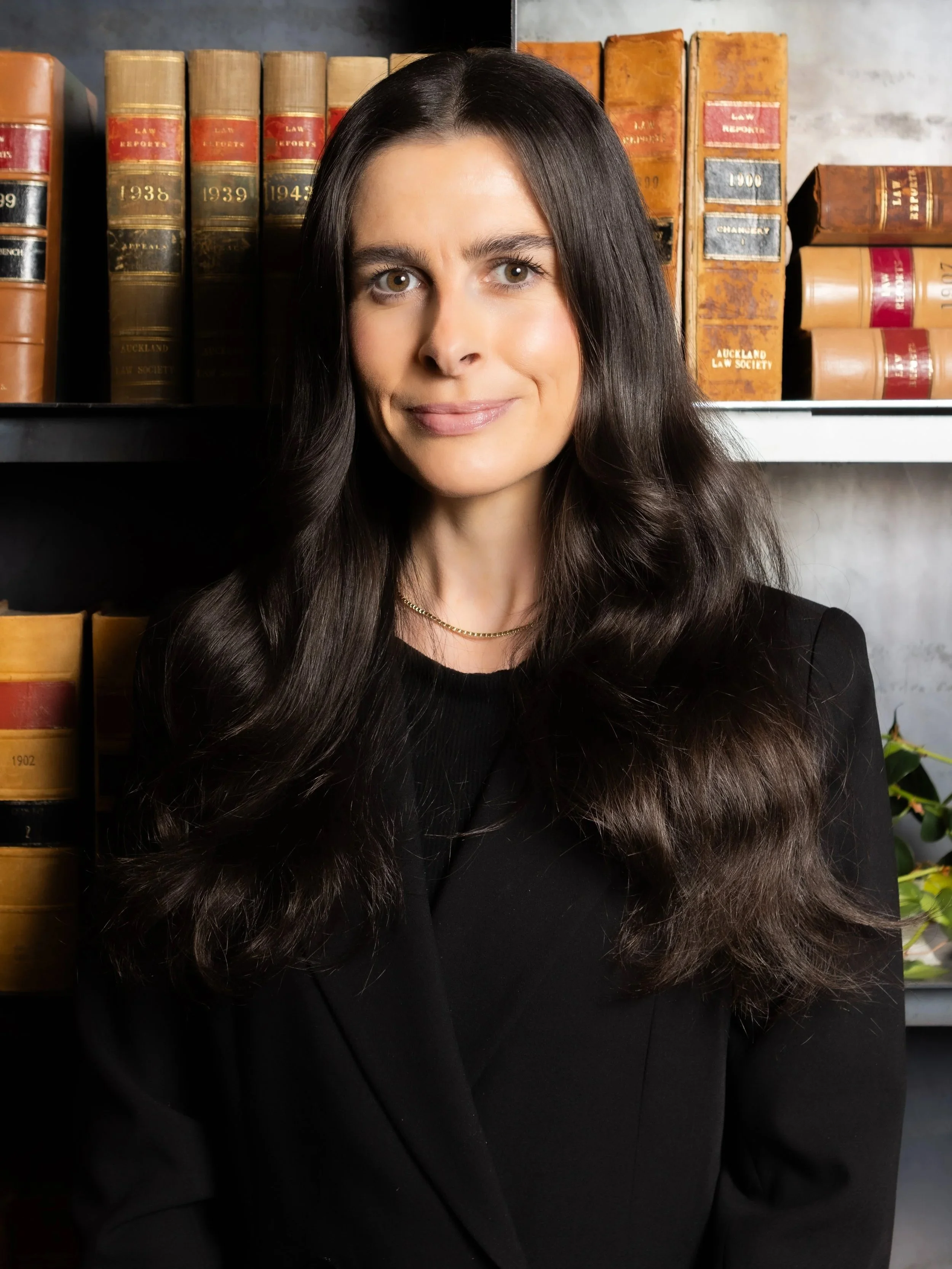 A woman with long dark hair wearing a black blazer and white blouse standing in front of a bookshelf filled with old books.