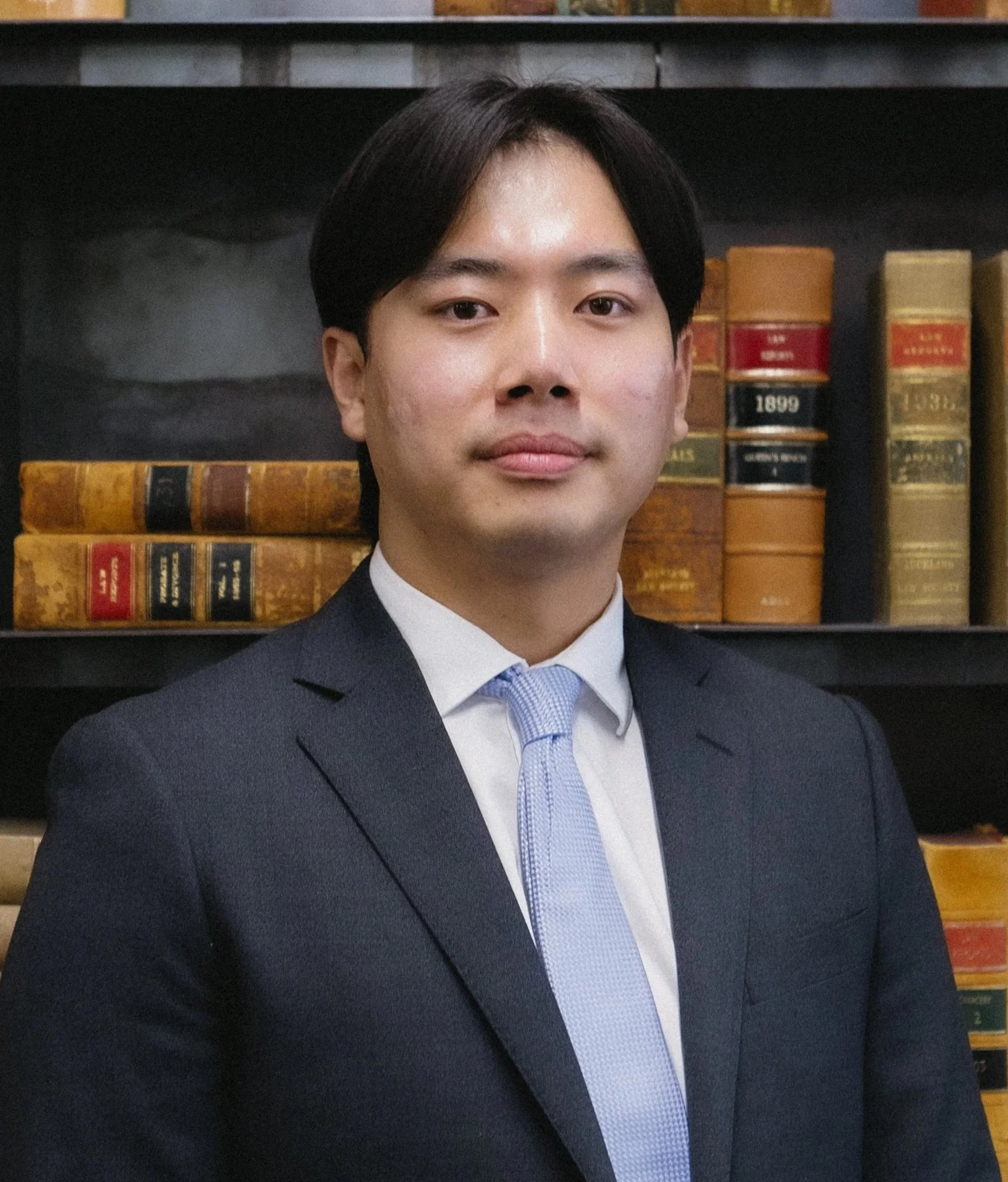 A young man in a suit and light blue tie standing in front of a bookshelf filled with old, leather-bound books.