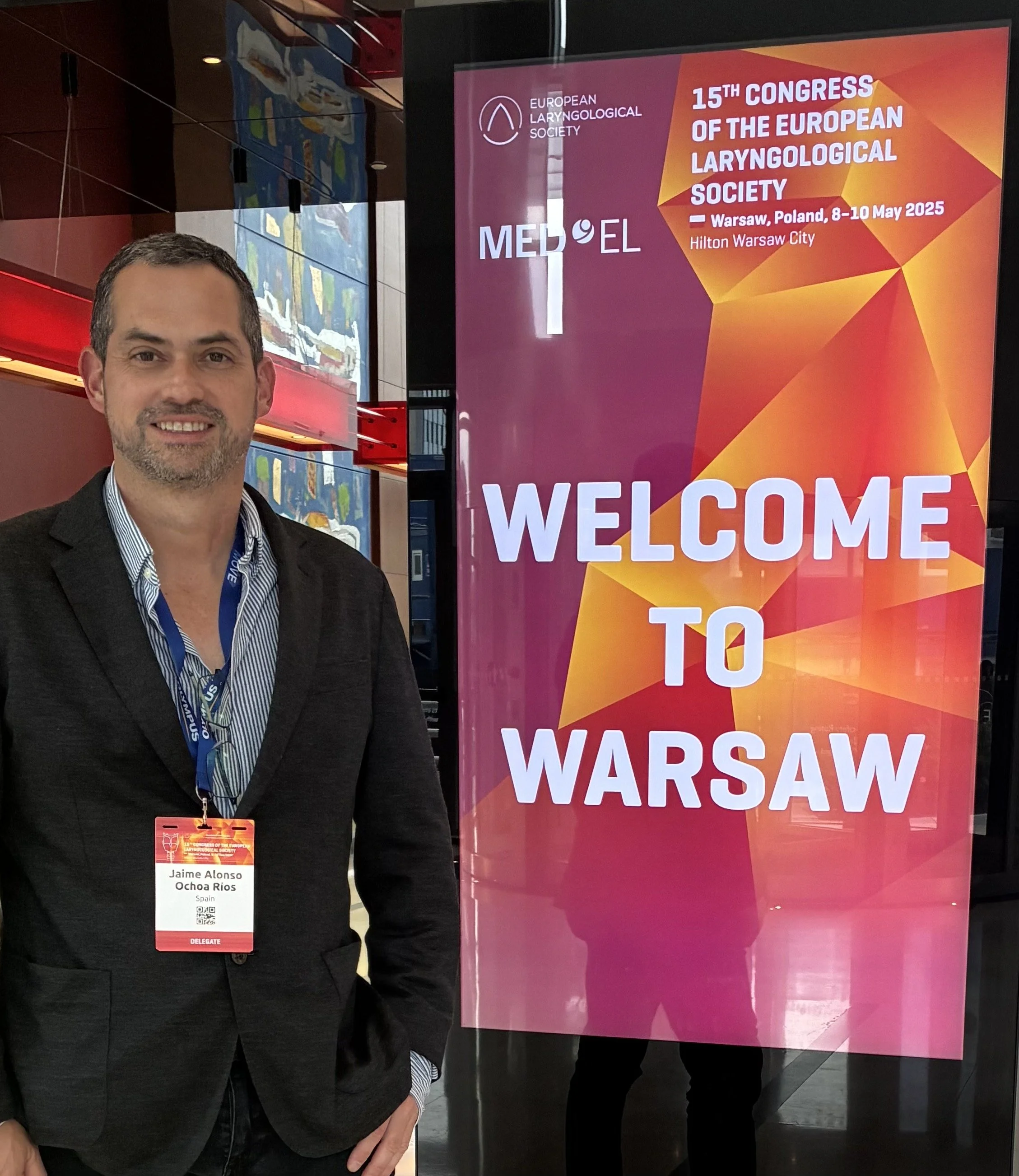 Hombre con una tarjeta de identificación en un evento, de pie junto a una cartelera que dice 'Welcome to Warsaw' en un congreso de laringología en Varsovia, Polonia, del 8 al 10 de mayo de 2025.