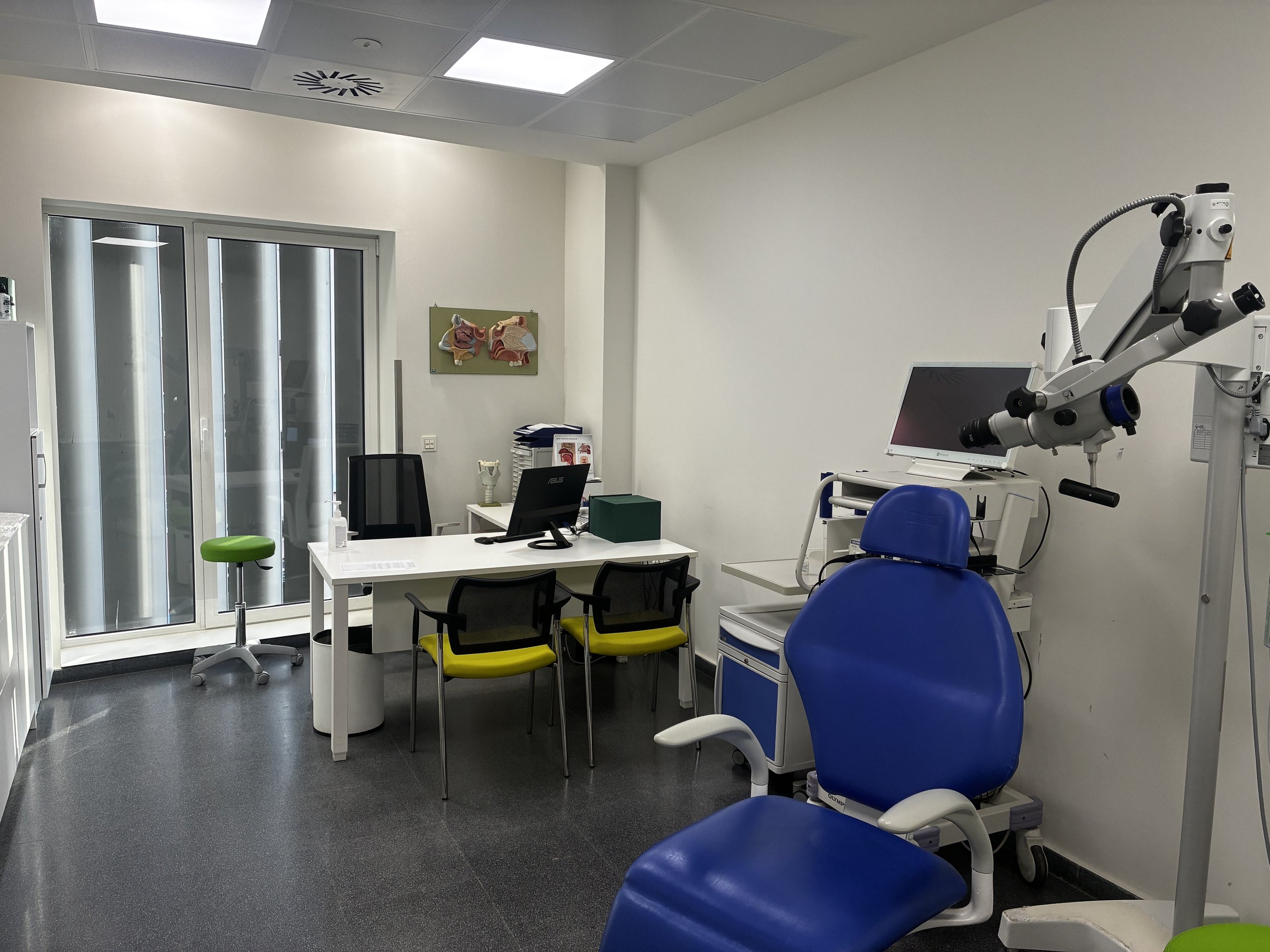Consultorio médico con equipo de diagnóstico, silla de examen, escritorio, computadoras y una ventana con persianas.