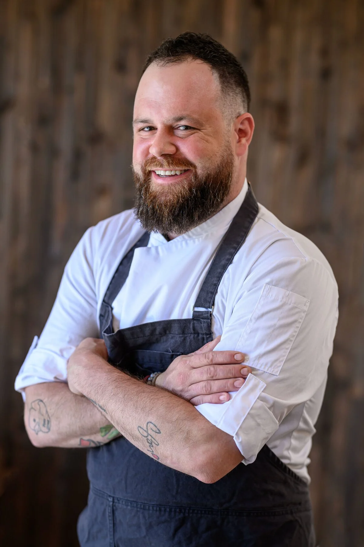 Executive Chef George Morris of The Hudson Bend with a beard and tattoos on his arms, wearing a white chef's coat and black apron, standing with arms crossed in front of a wooden background.