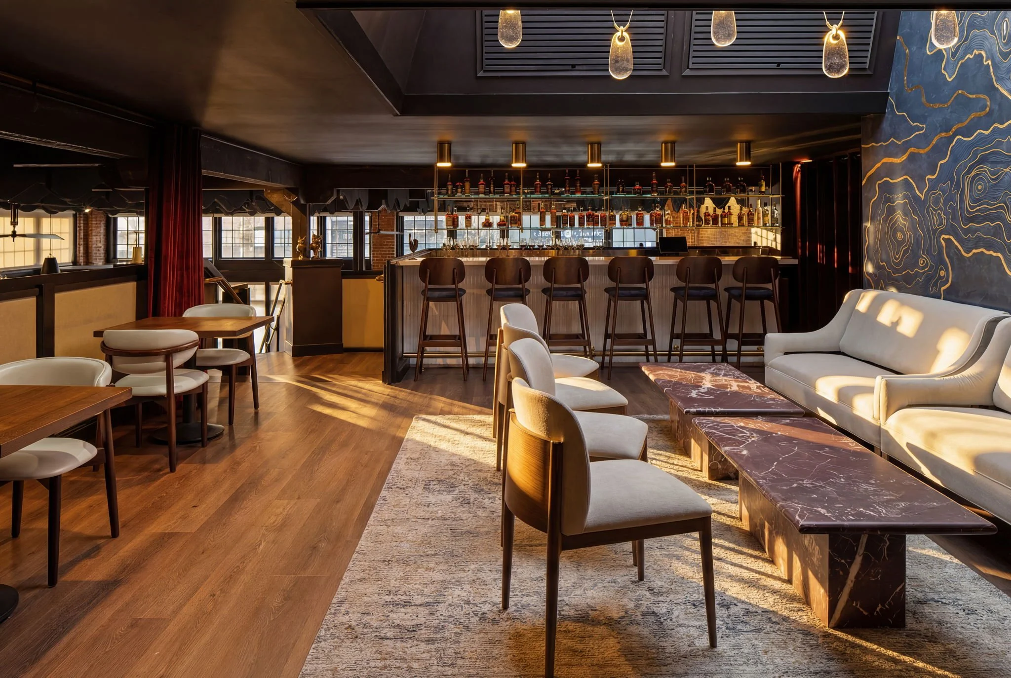 Interior of a modern bar with a wooden bar counter, bar stools, and a wall decorated with a large abstract mural, illuminated by warm hanging lights.