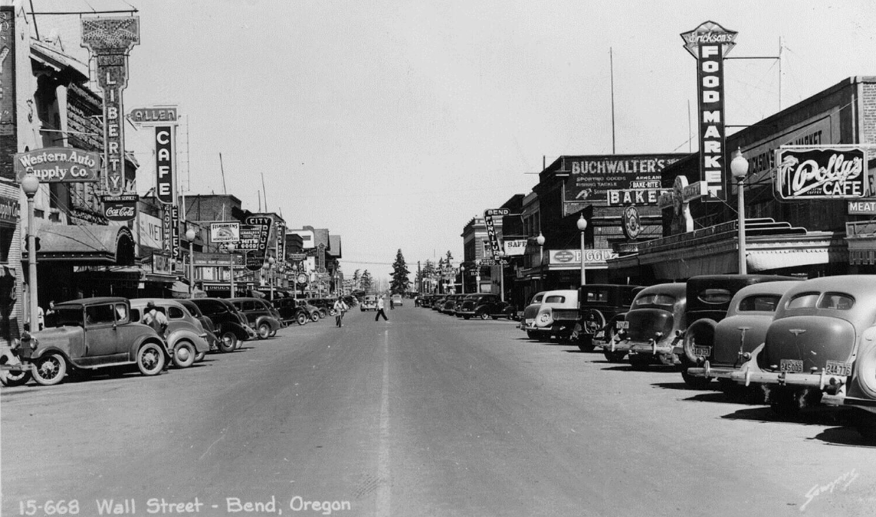 A black and white vintage street scene of Wall Street in Bend, Oregon, showing parked cars lining both sides of the street and various storefronts with large signs, including a food market, bakery, cafe, and auto supply shop. Pedestrians are walking along the sidewalk.