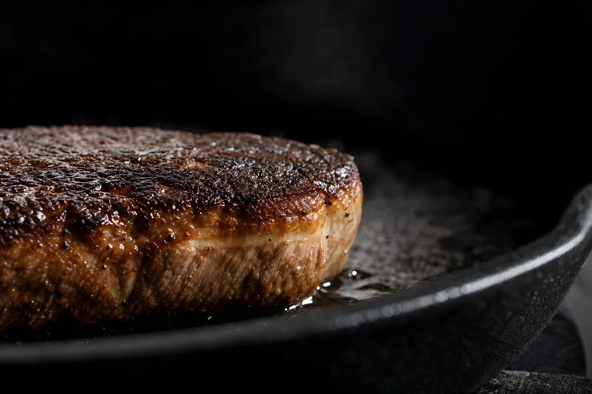 Close-up of a cooked, seasoned steak frying on a black skillet.
