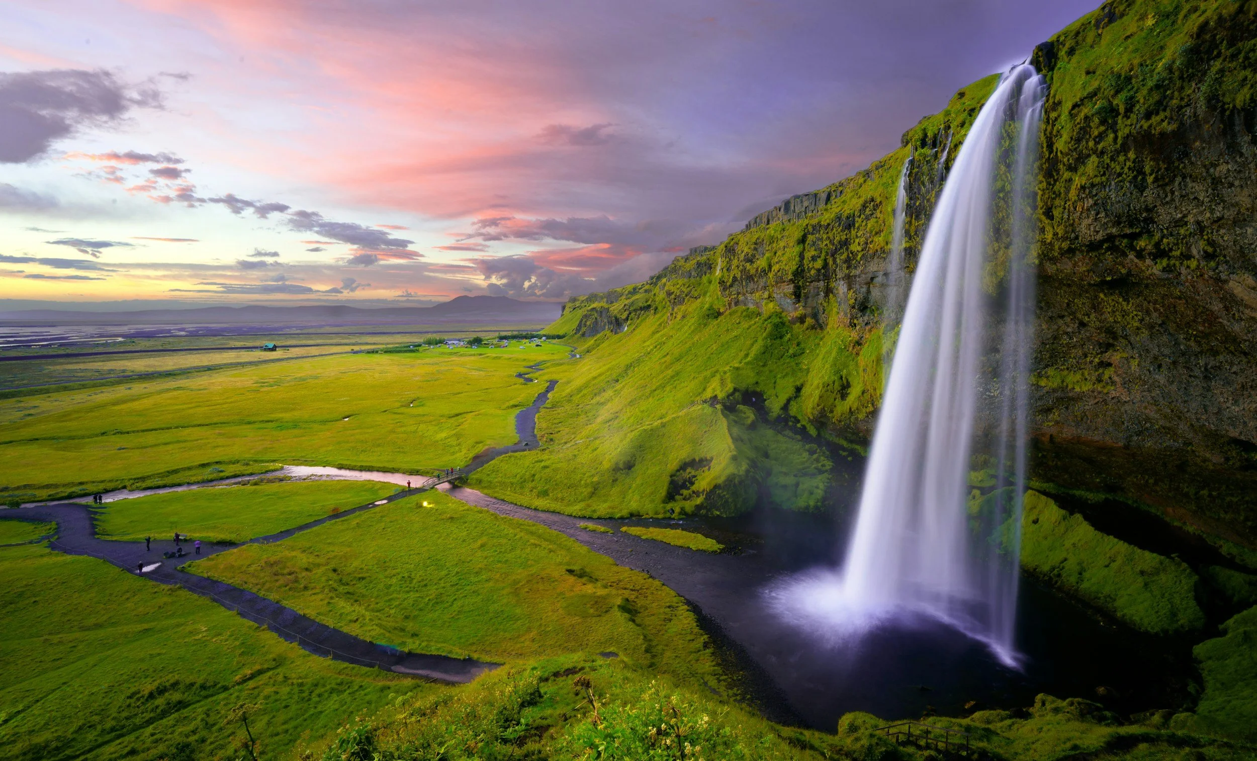 A scenic landscape of a waterfall flowing down a moss-covered cliff into a pond, with lush green fields, a winding path, and a colorful sky at sunset.