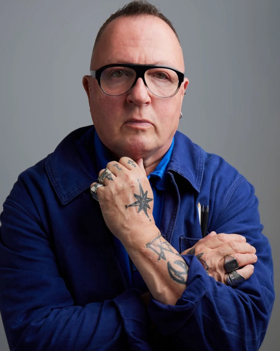 Serge facing the camera wearing glasses, hand tattoos, and silver rings poseing with hand on chin against a plain background.