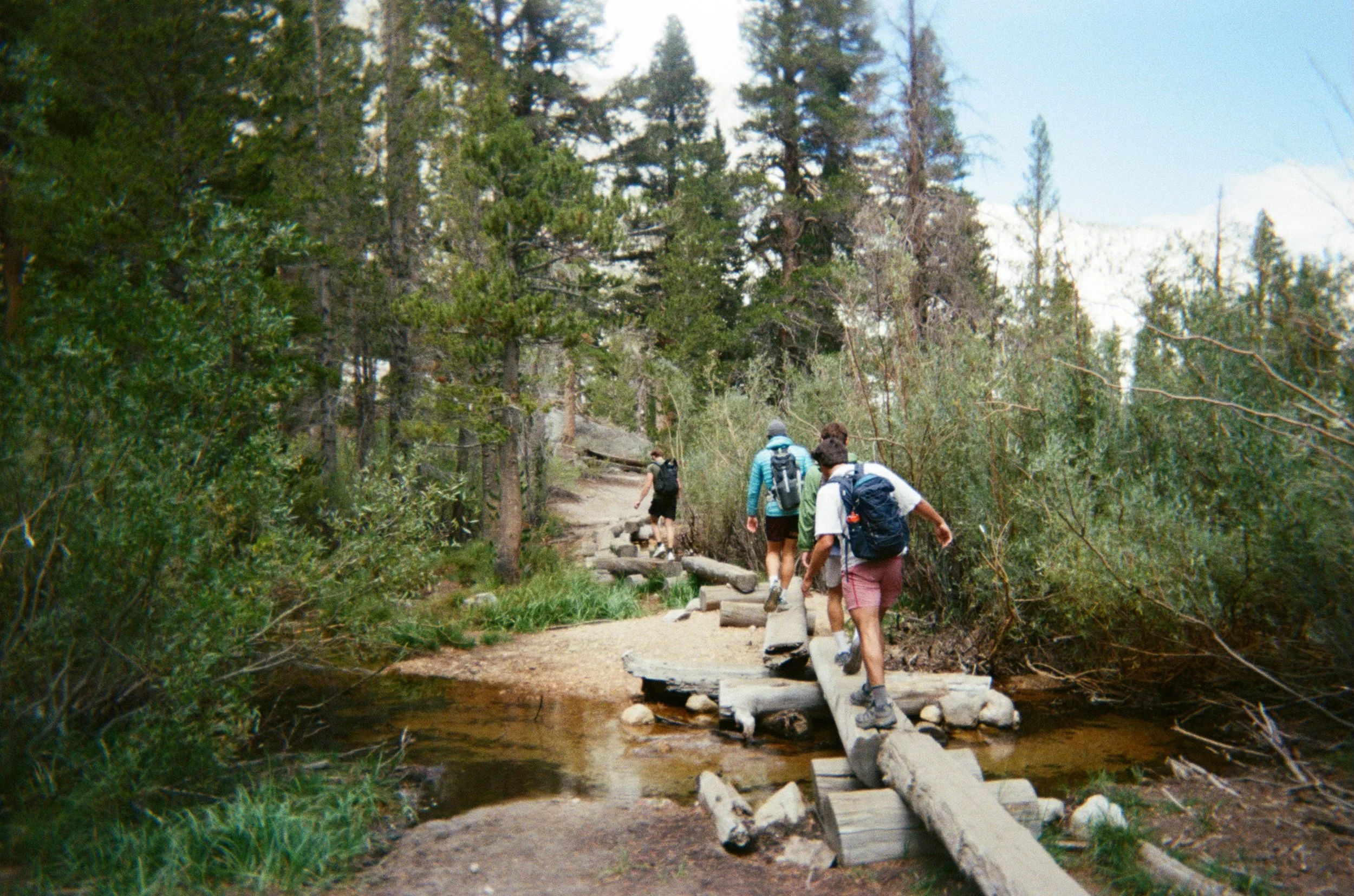 Group of hikers crossing a narrow wooden bridge over a creek in a forested mountain trail.