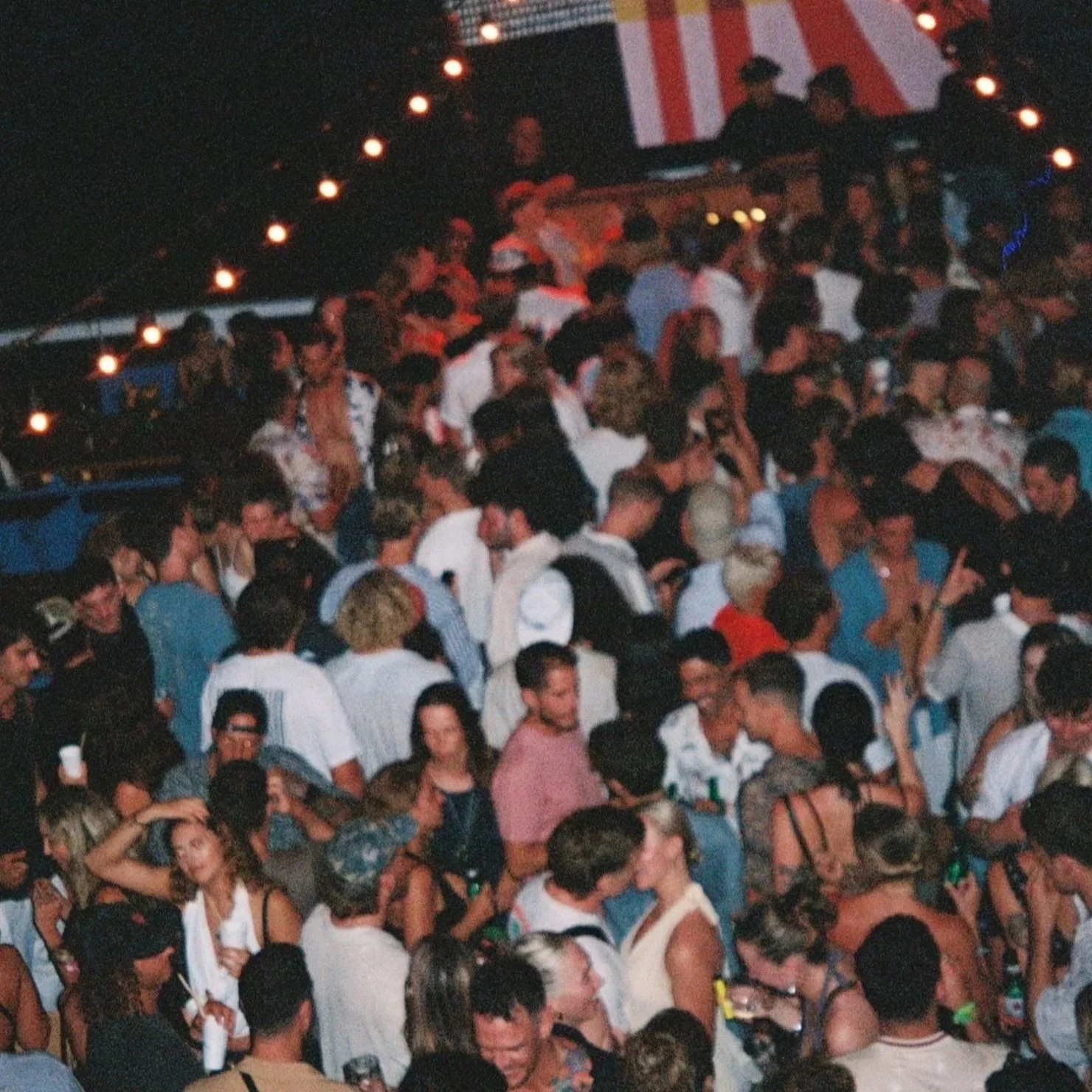 Crowd of people at a party or concert, some holding drinks, illuminated by string lights at night.