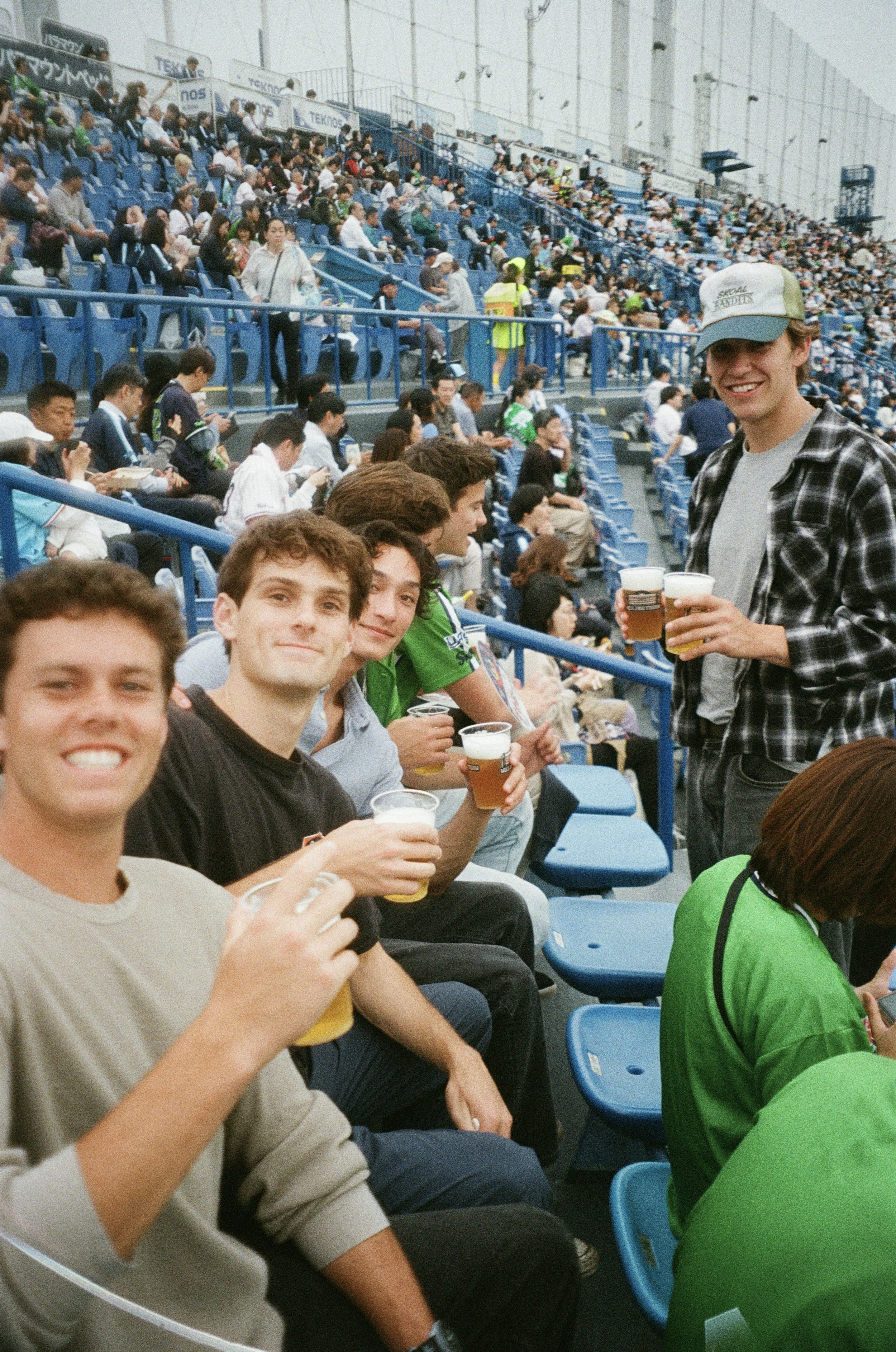 Group of young people sitting in stadium seats drinking beer and smiling on a cloudy day.