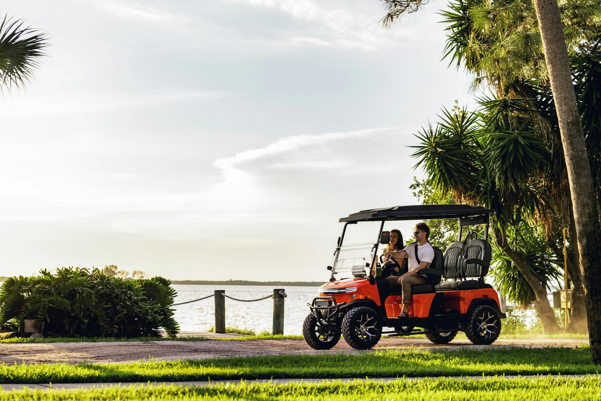 Couple riding in a red electric golf cart along a scenic lakeside path with lush palm trees, bushes, and a calm lake in the background during daytime.