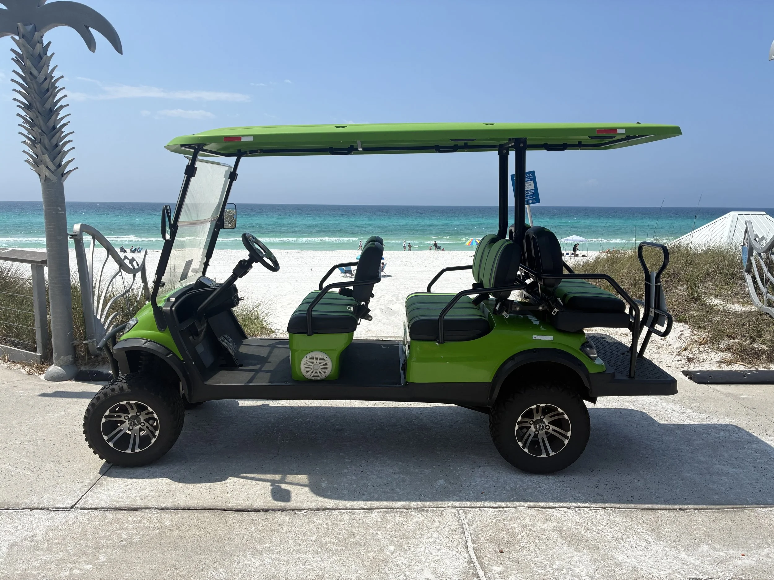 Couple riding in a red electric golf cart along a scenic lakeside path with lush palm trees, bushes, and a calm lake in the background during daytime.