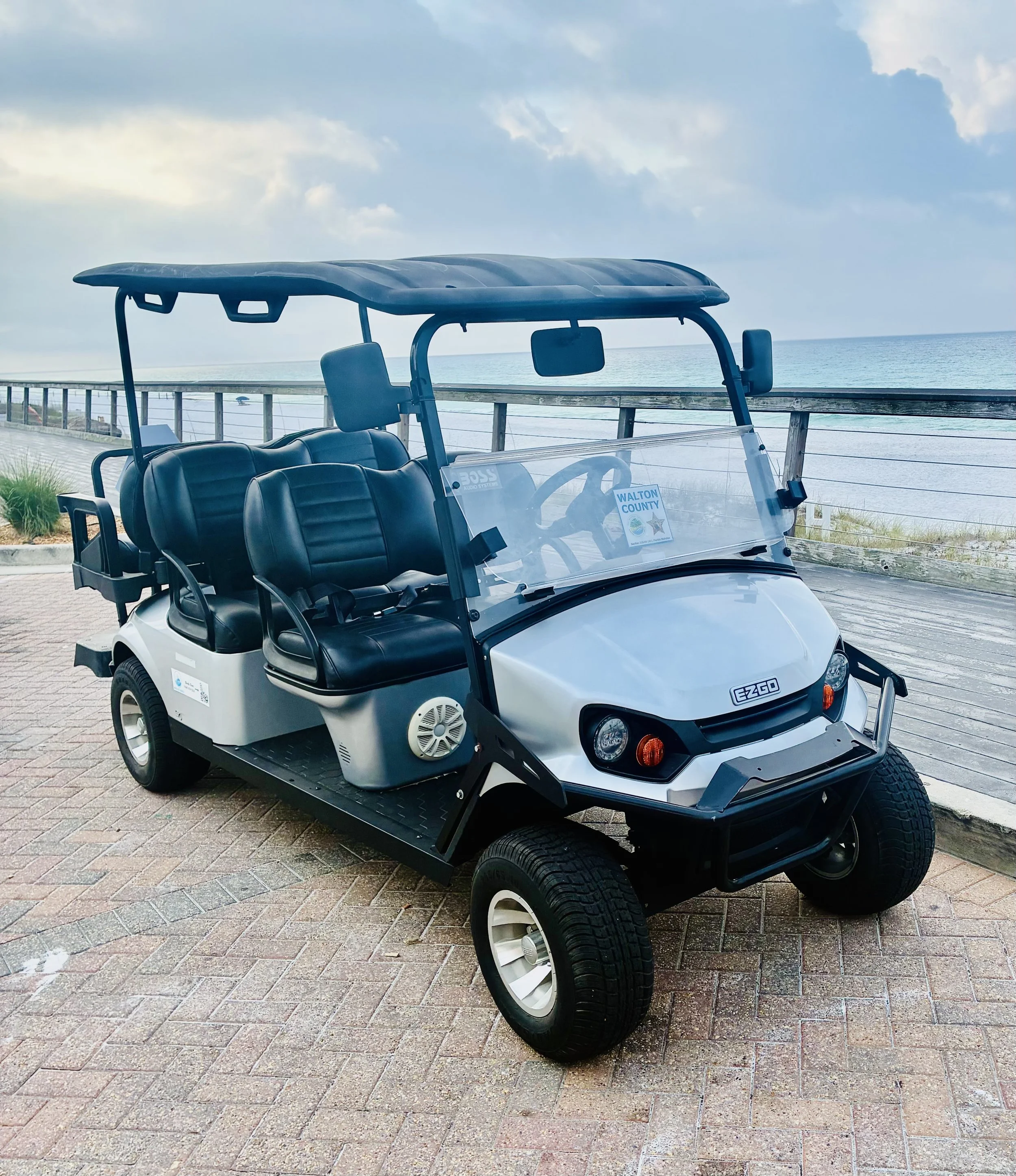 A red golf cart parked on a beachside path with multiple tan seats, a clear windscreen, and a canopy overhead, with the ocean and sky in the background.
