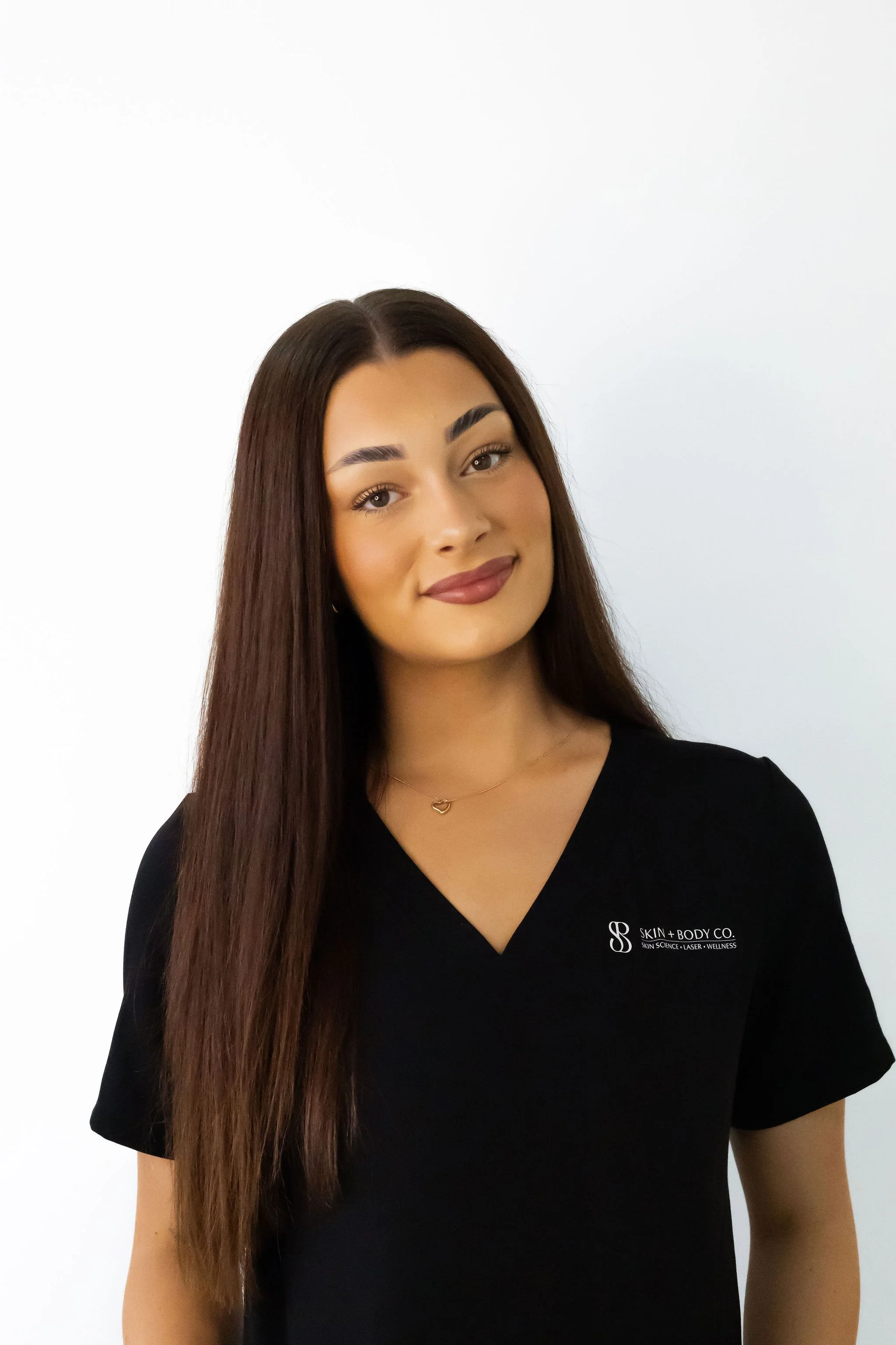 A young woman with long brown hair, wearing a black V-neck shirt with a logo that reads 'Skin + Body Co.'. She has light skin, makeup, and is smiling slightly, standing against a plain white background.