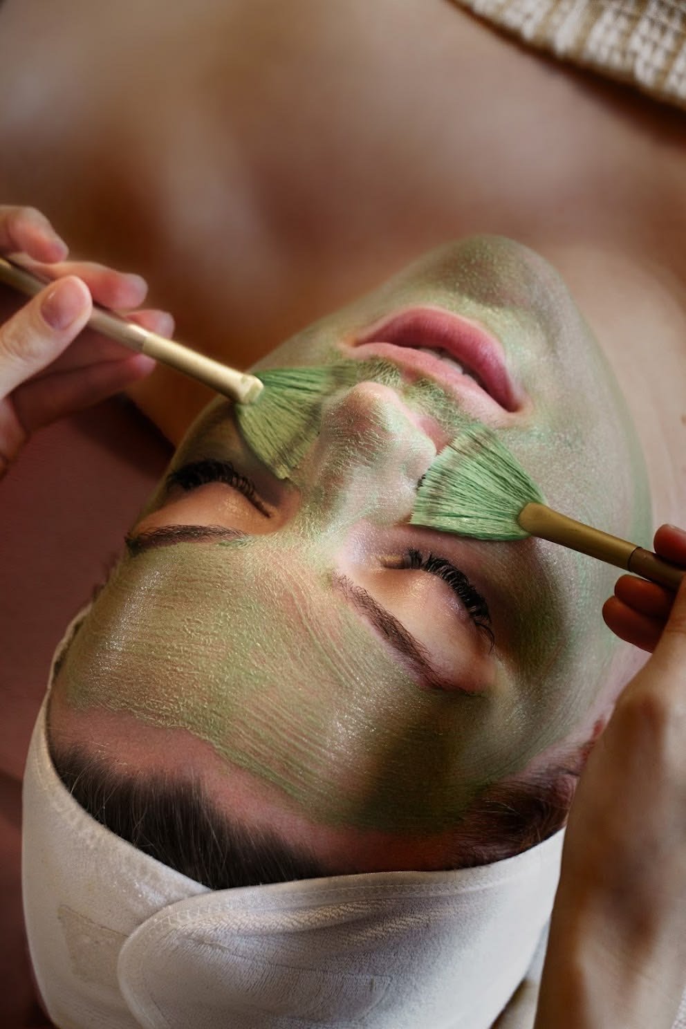 Woman receiving a facial treatment with green face mask application using two brushes.